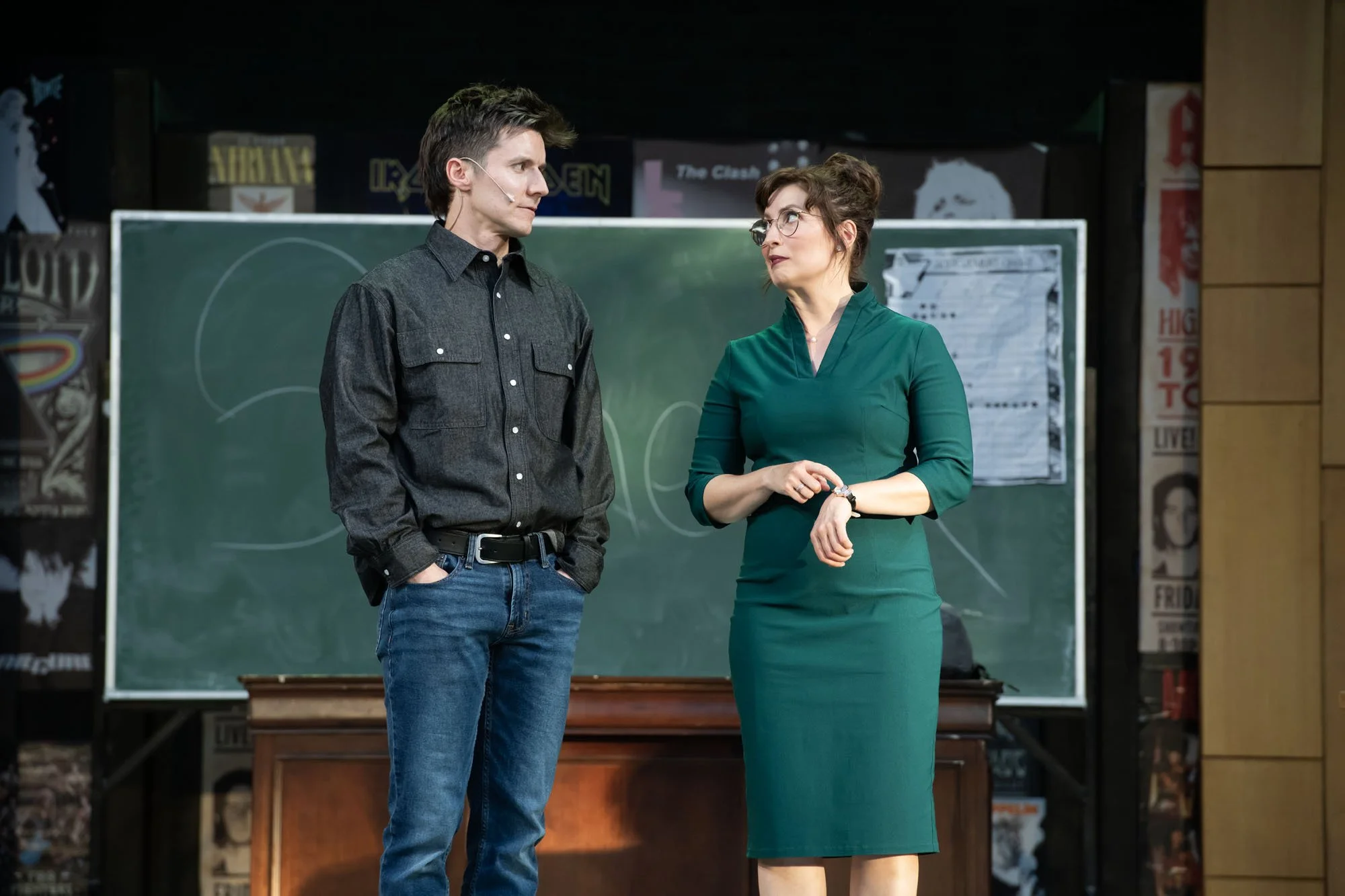 A man and woman standing in front of a blackboard in a classroom, engaging in conversation.