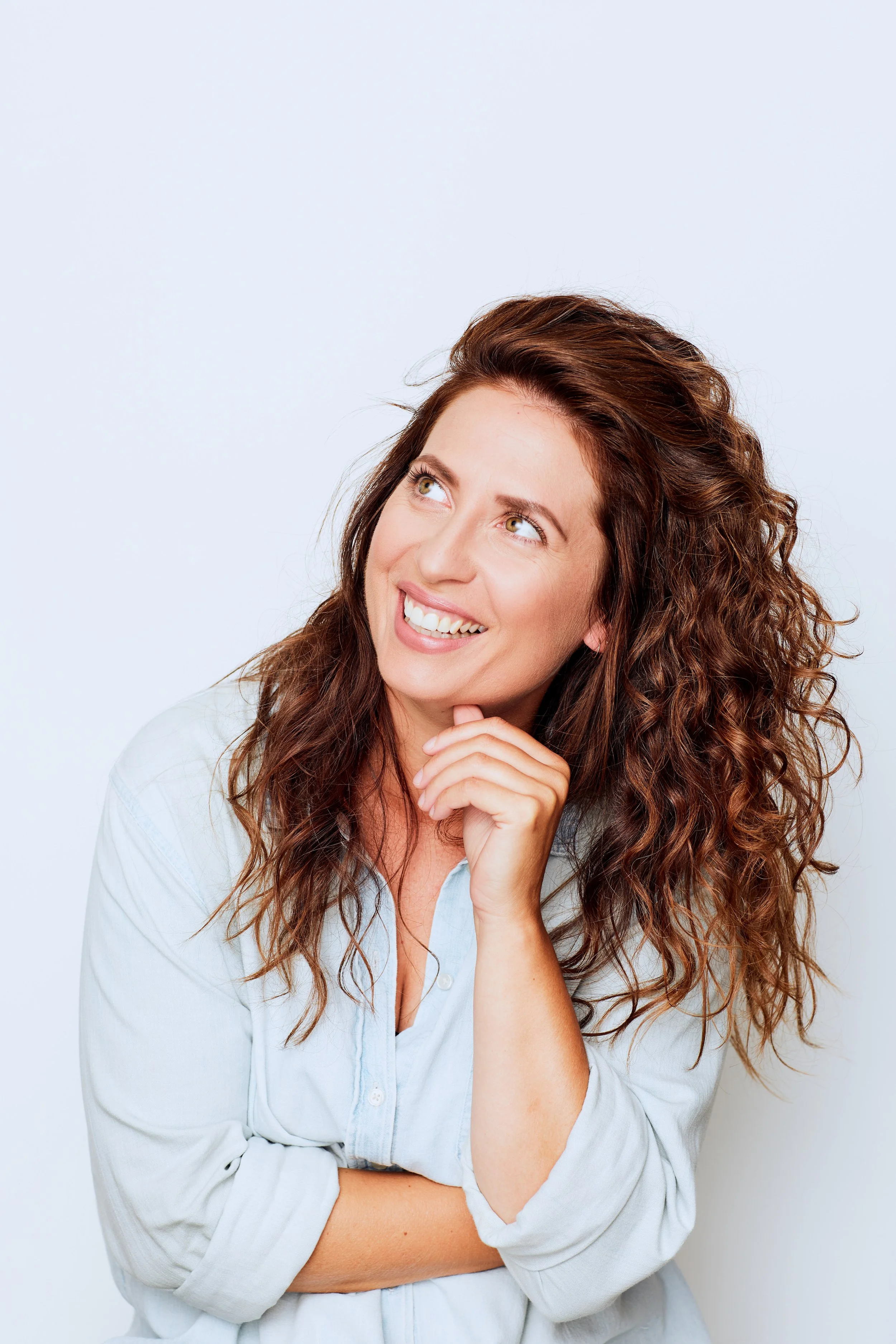 Woman with curly brown hair smiling and looking up, wearing a light blue button-down shirt, against a plain white background.