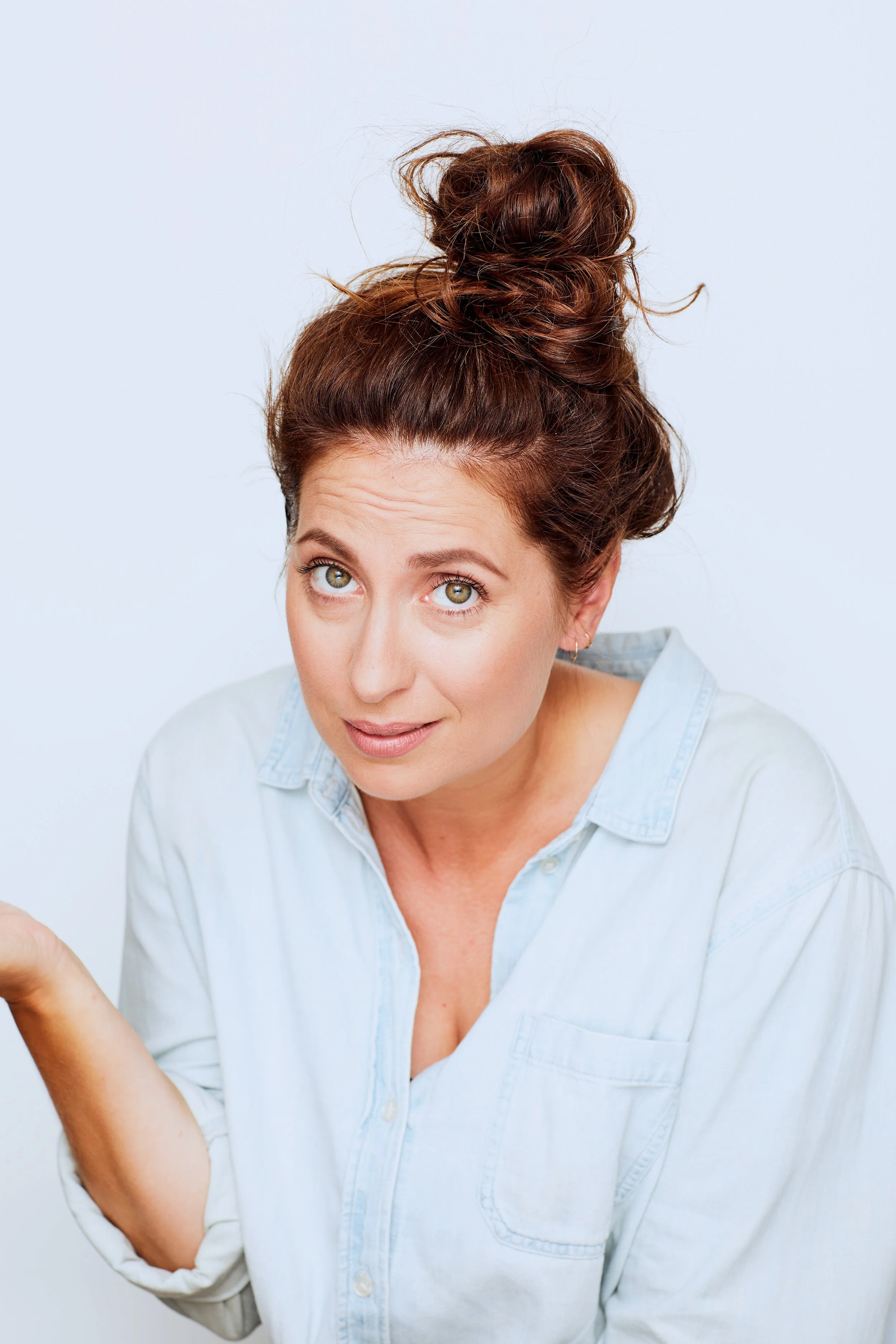 A woman with light skin, brown hair tied in a messy bun, and wearing a light-colored button-up shirt raises her eyebrows and looks at the camera with a slightly puzzled or inquisitive expression.