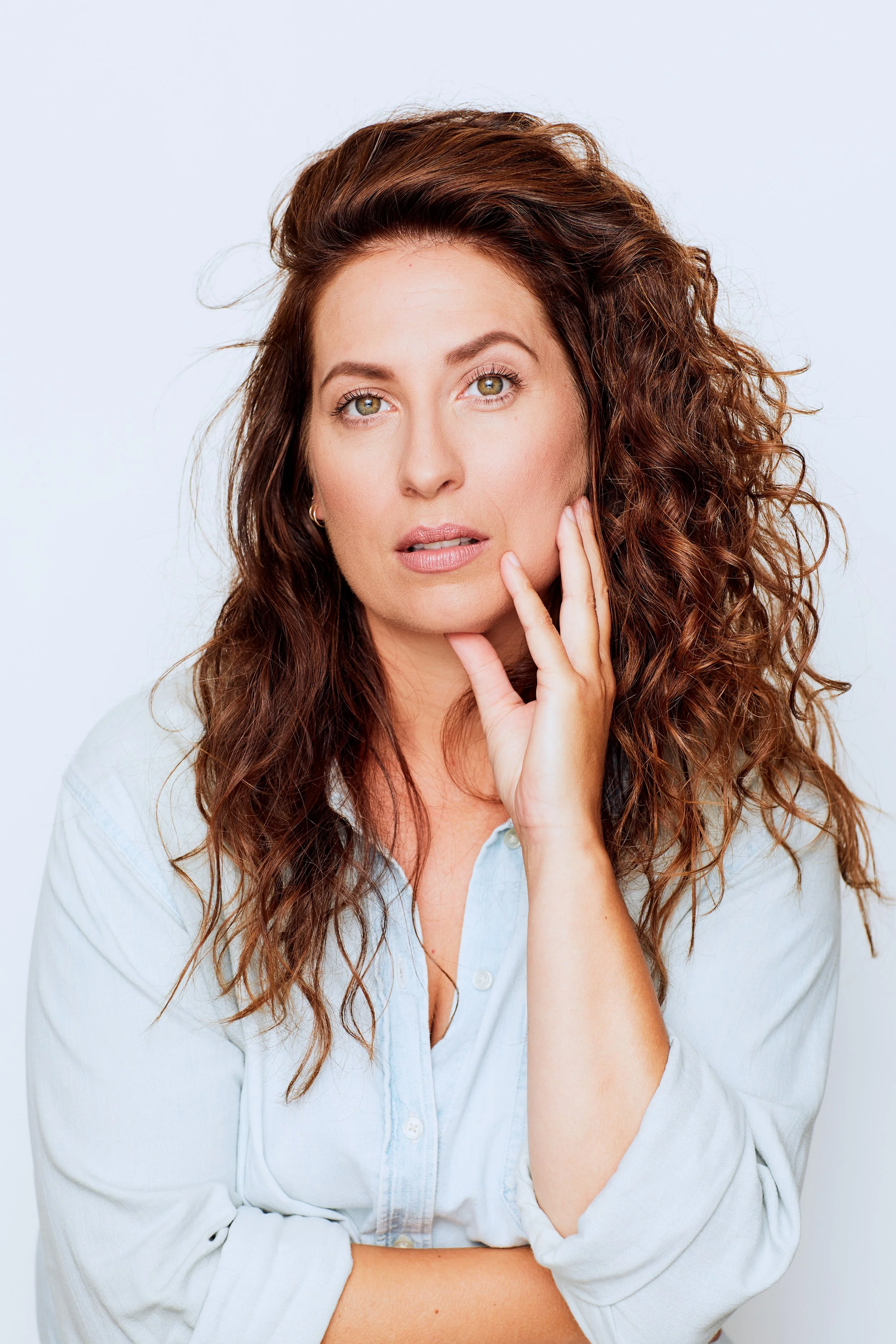 A woman with curly brown hair and light skin, wearing a light blue shirt, touching her face with her right hand, looking directly at the camera against a plain white background.