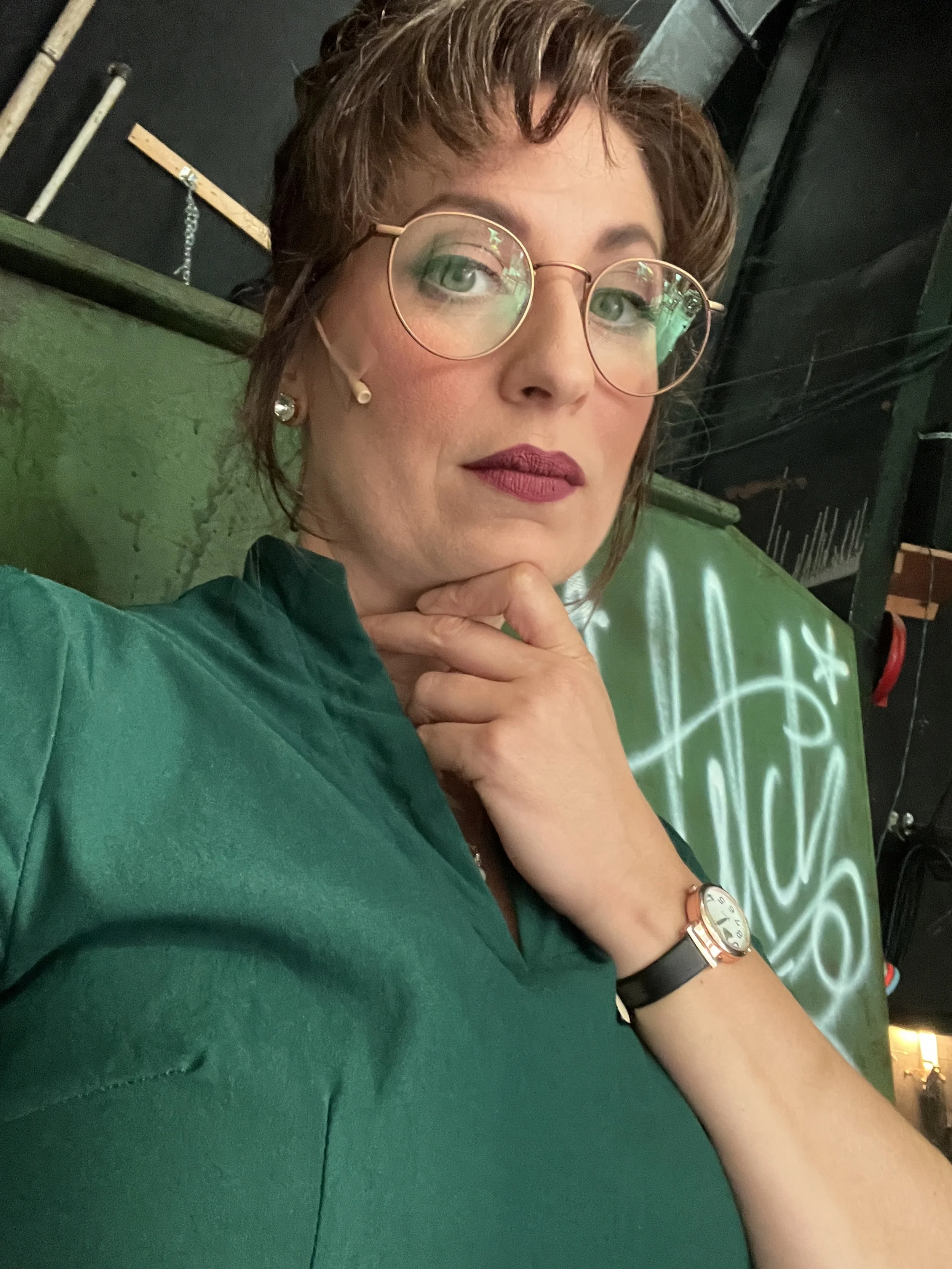 A woman with short curly hair and glasses poses with her hand touching her chin, wearing a green top and a watch, in front of a dark background with a green banner.