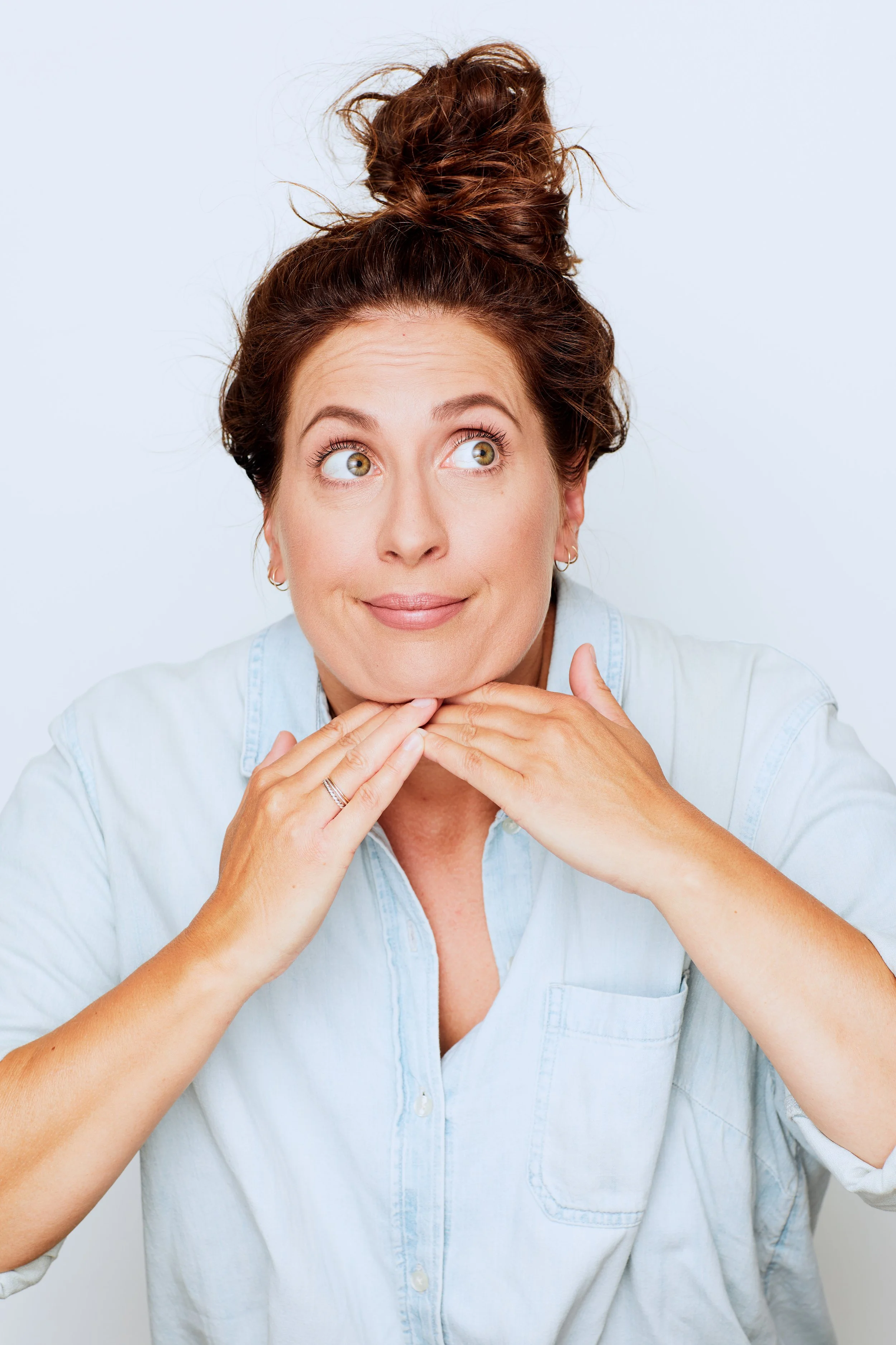 A woman with curly brown hair styled in a high messy bun is looking up and to the side with a thoughtful expression, wearing a light blue denim shirt.