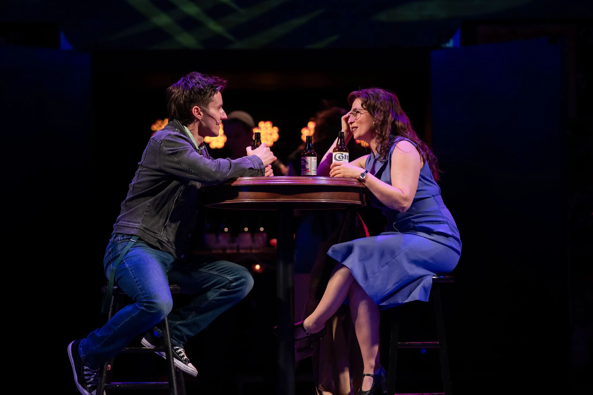 Two people, a man and a woman, sit at a round table on a stage in a dimly lit setting, having a conversation and smiling, with bottles of beer in front of them.