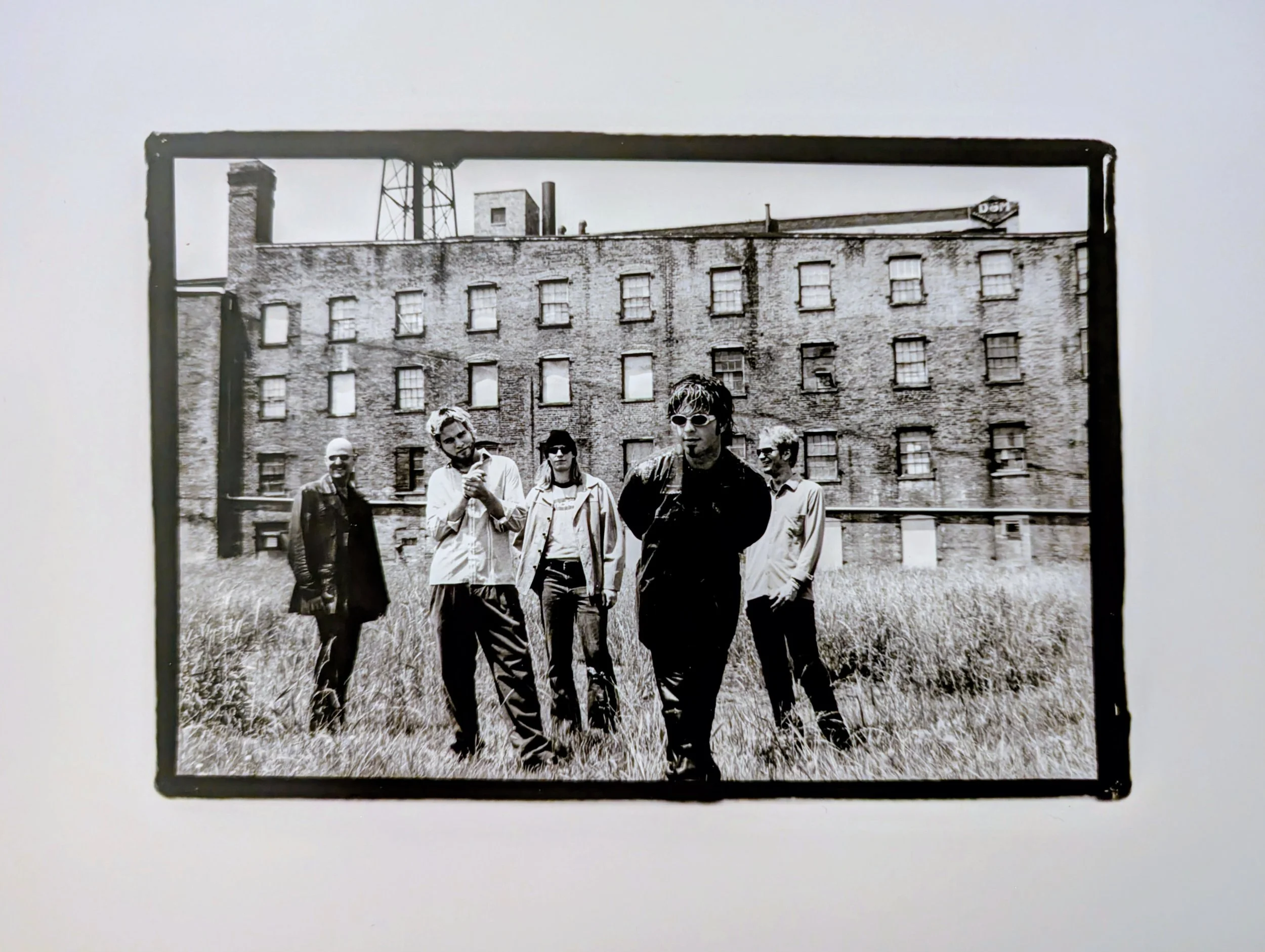 Black and white photo of five people standing outdoors in front of a large, run-down brick building with multiple windows, some of which are boarded up, and tall grass in the foreground.