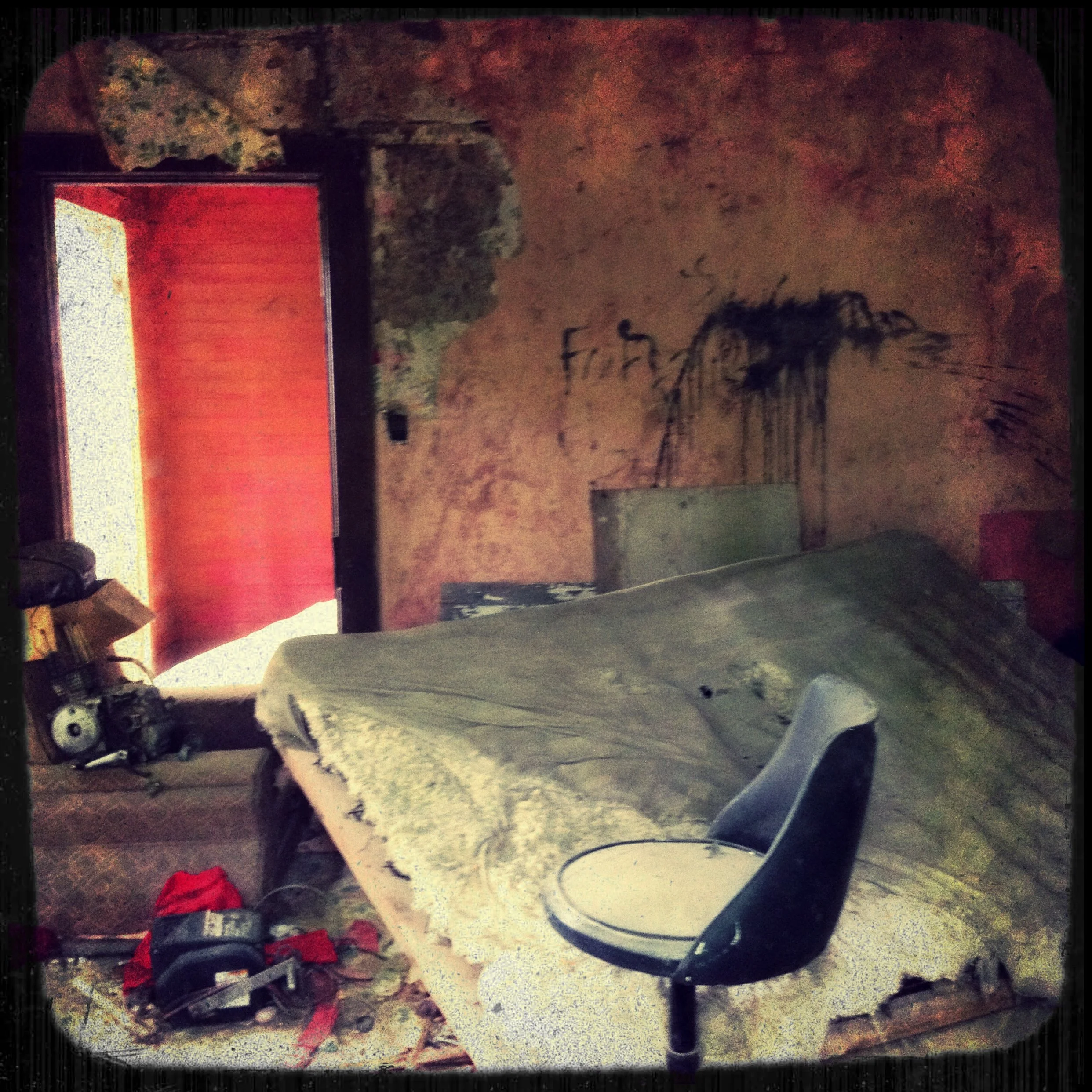 A cluttered, dilapidated bedroom with a bed covered in dirty sheets, a broken chair, and various tools and debris on the floor. The wall has peeling paint and graffiti, and a window with a red curtain partially open.