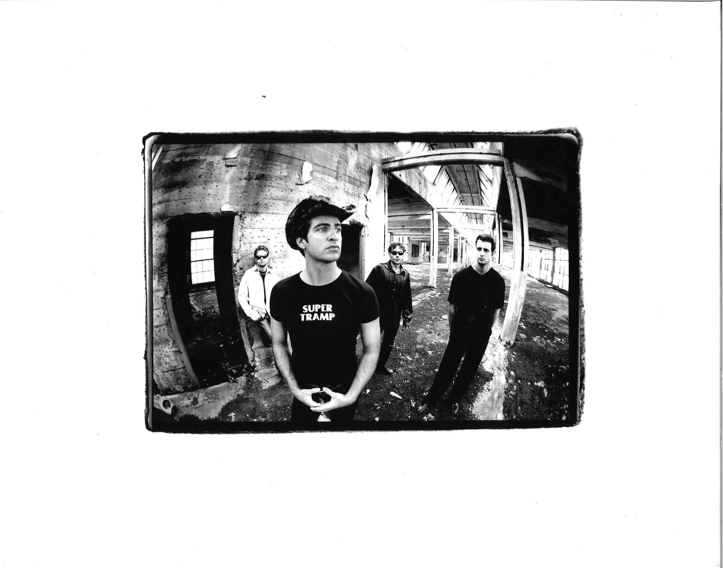 Black and white photo of four young men inside an abandoned building with large windows and exposed wooden beams. One in the front wears a shirt that says 'Super Tramp'.