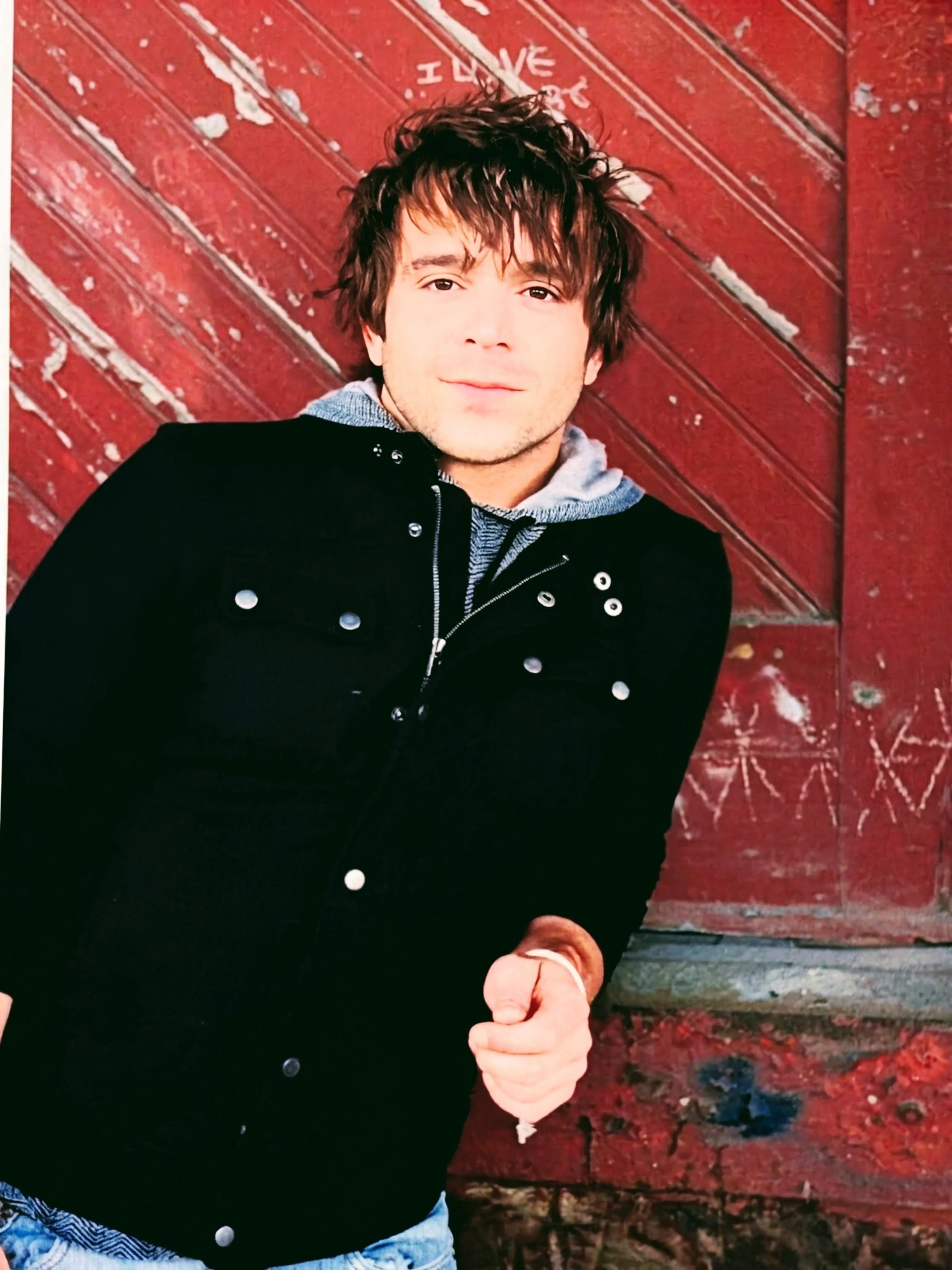 Young man with dark, tousled hair wearing a black jacket over a gray hoodie, standing against a weathered red barn door with chalk drawings.