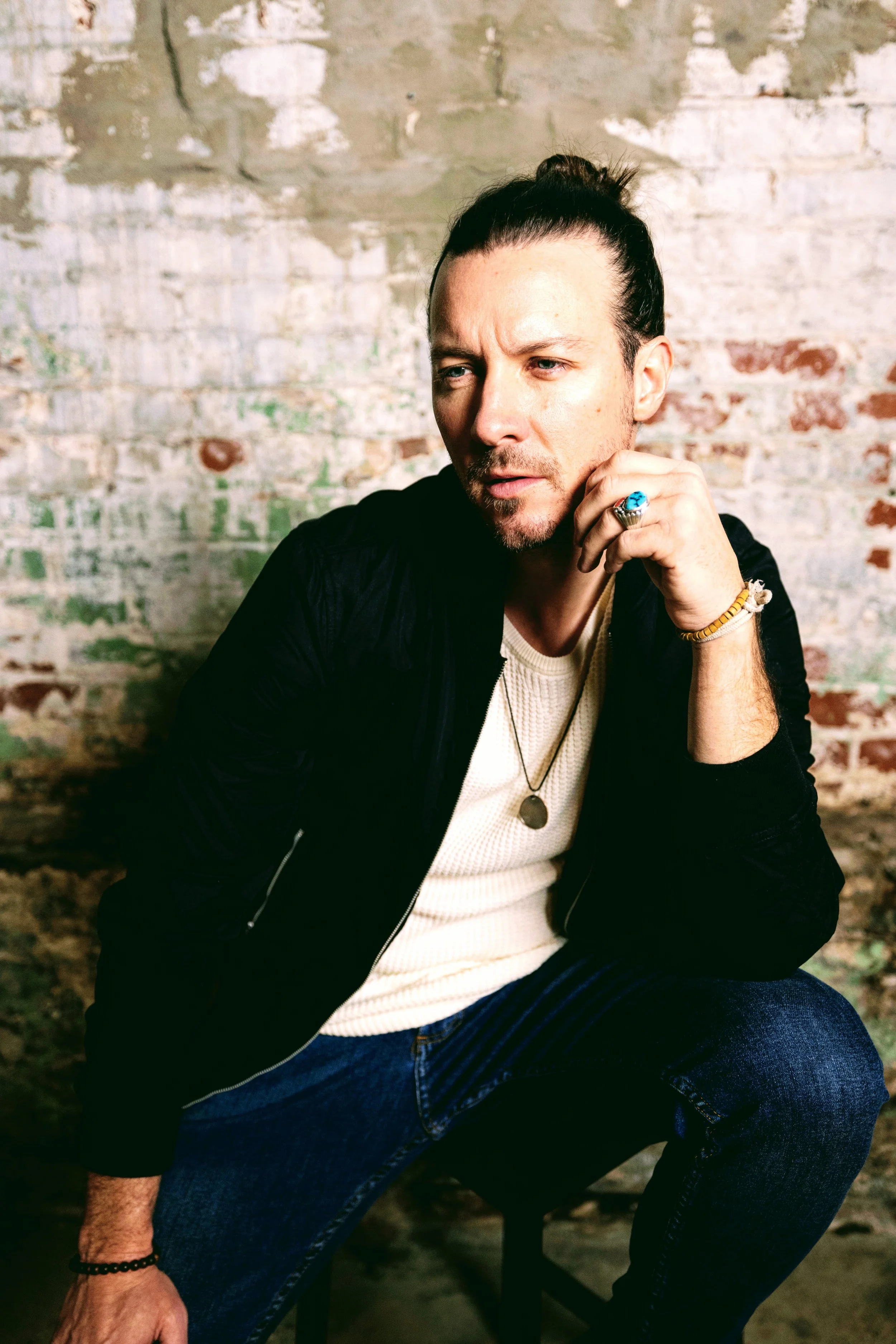 A man with long dark hair tied back, wearing a black jacket, cream shirt, and jeans, sitting against a weathered brick wall with a contemplative expression.