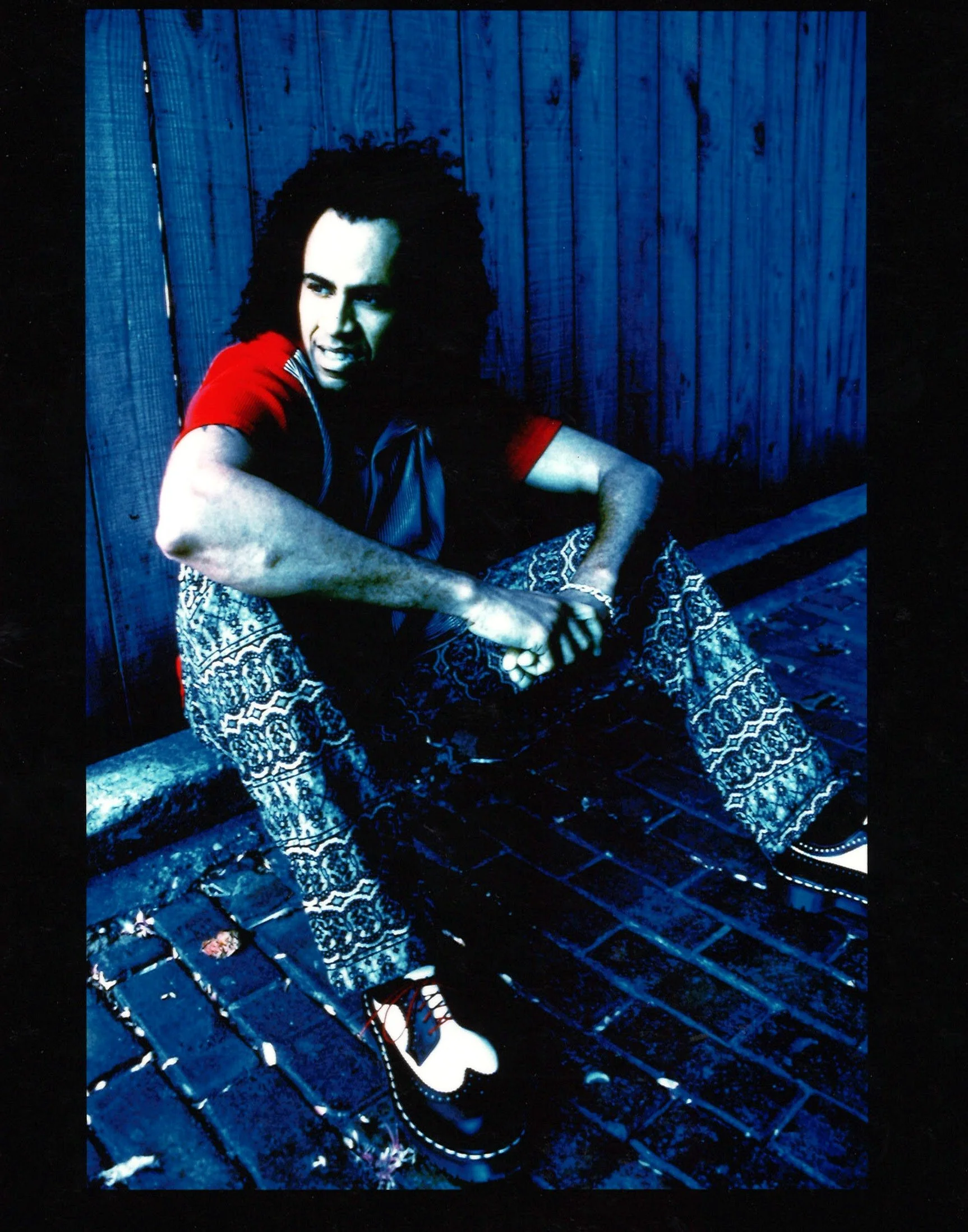 A person with dark curly hair sitting on a brick sidewalk, leaning against a wooden fence, wearing patterned pants, black and white shoes, a red and black shirt, with an expressive facial expression.