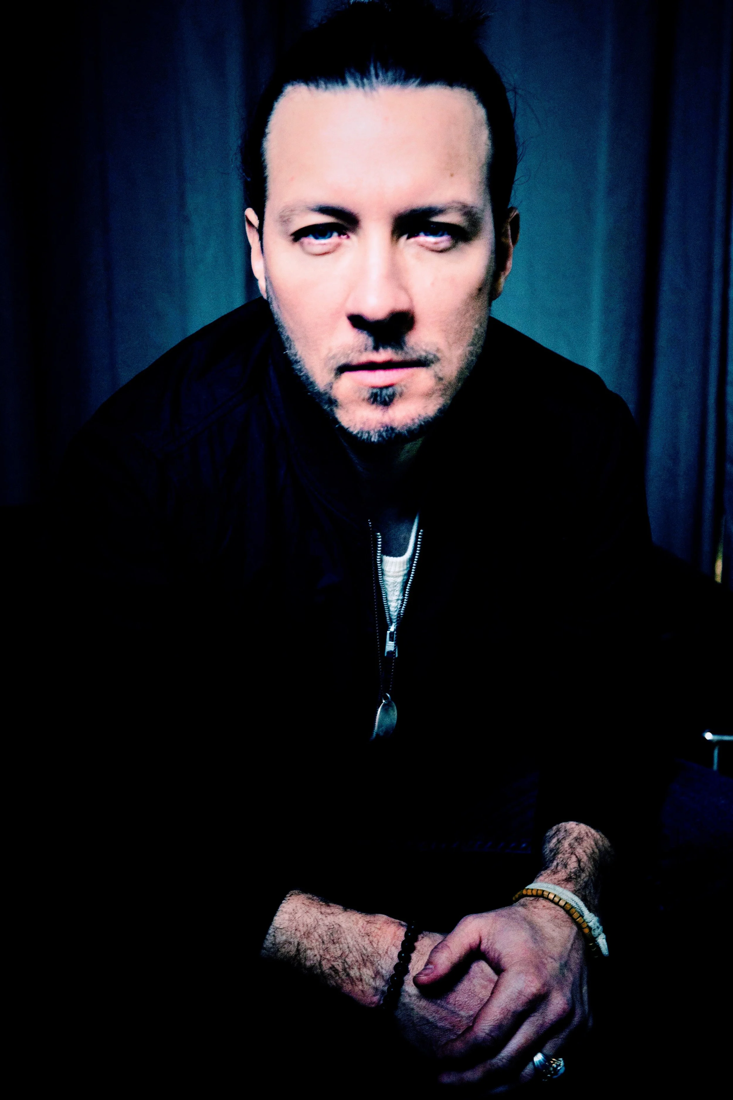 A man with slicked-back hair, wearing a black jacket with a zipper, sitting with his hands clasped, facing forward with a serious expression, in front of a dark blue background.