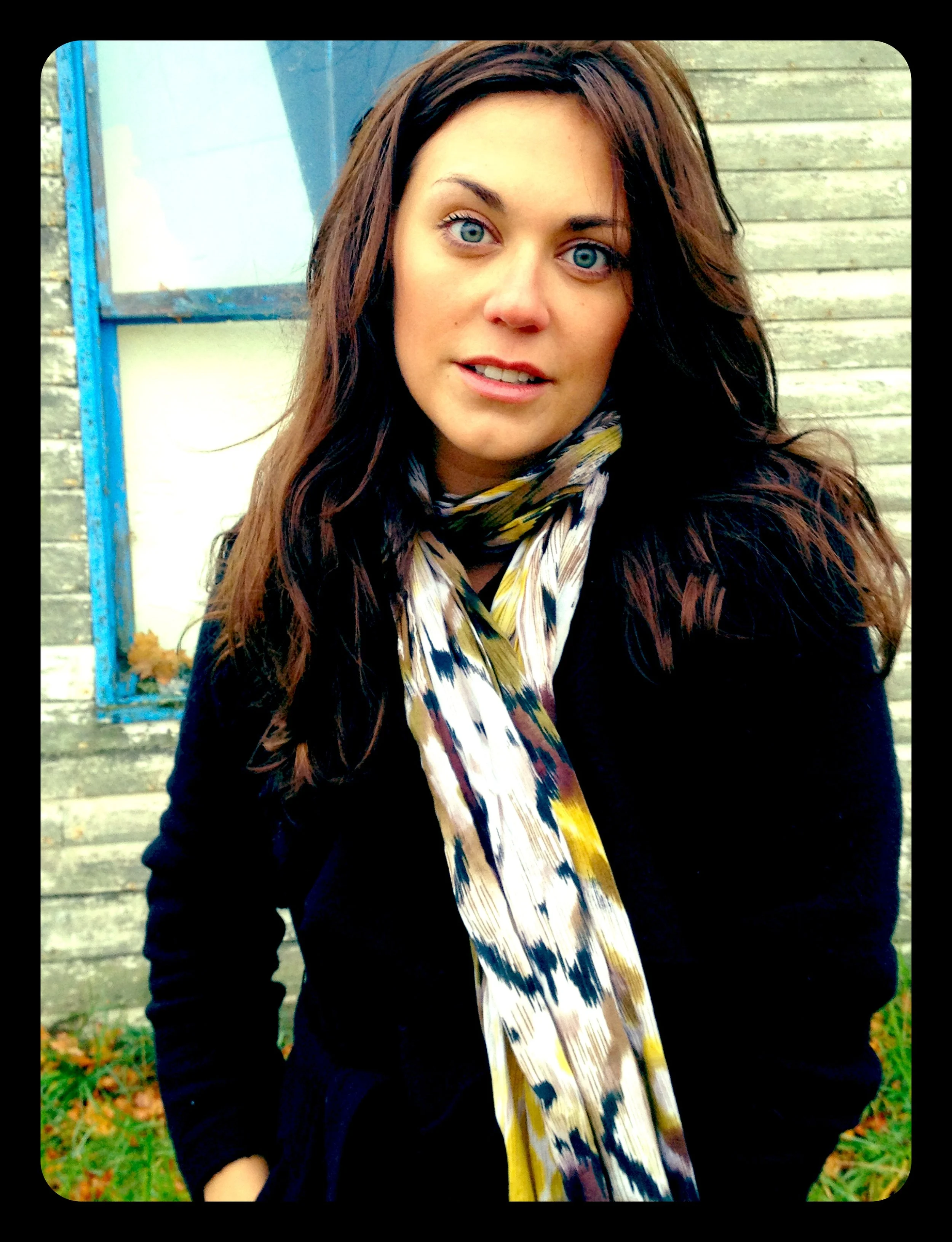 A woman with blue eyes and long brown hair standing outside in front of a weathered wooden wall and a window with a blue frame, wearing a dark jacket and a patterned scarf.