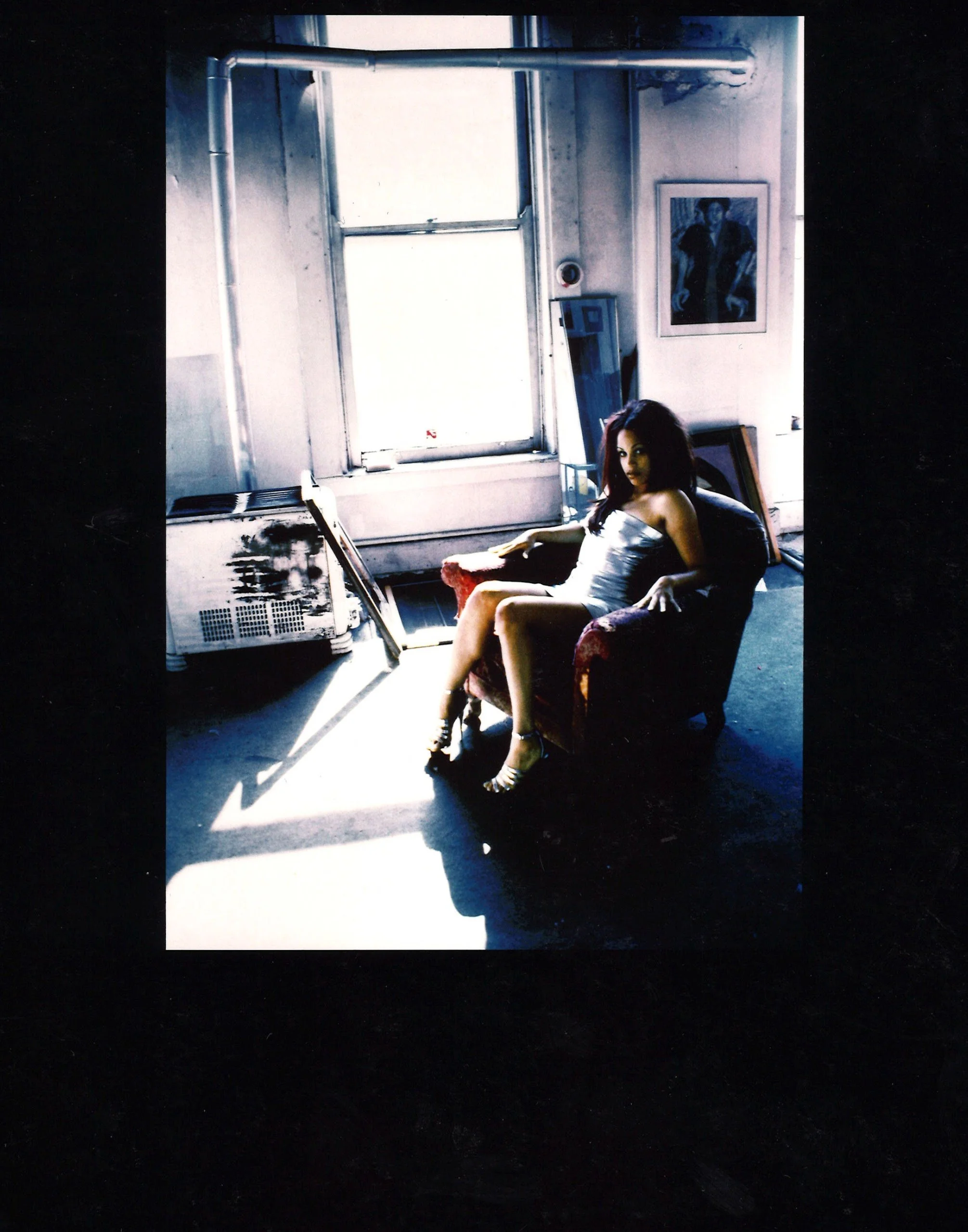 A woman in a silver dress sitting on a red armchair in a bright room with a large window, framed artwork on the wall, and paintings leaning against the wall.