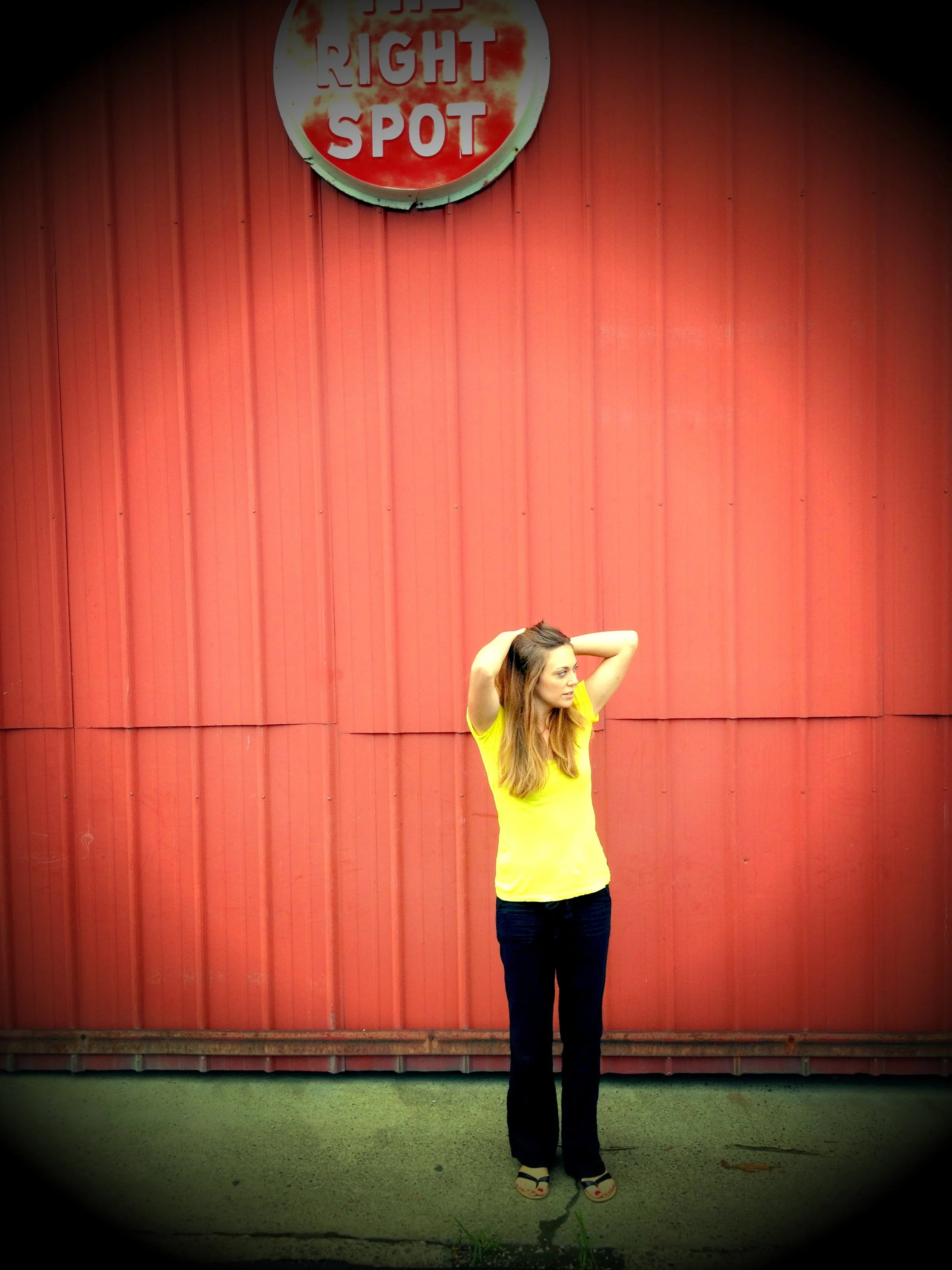 A woman in a yellow shirt and black pants standing with her arms raised behind her head in front of a red metal wall with a circular sign that reads 'Right Spot'.