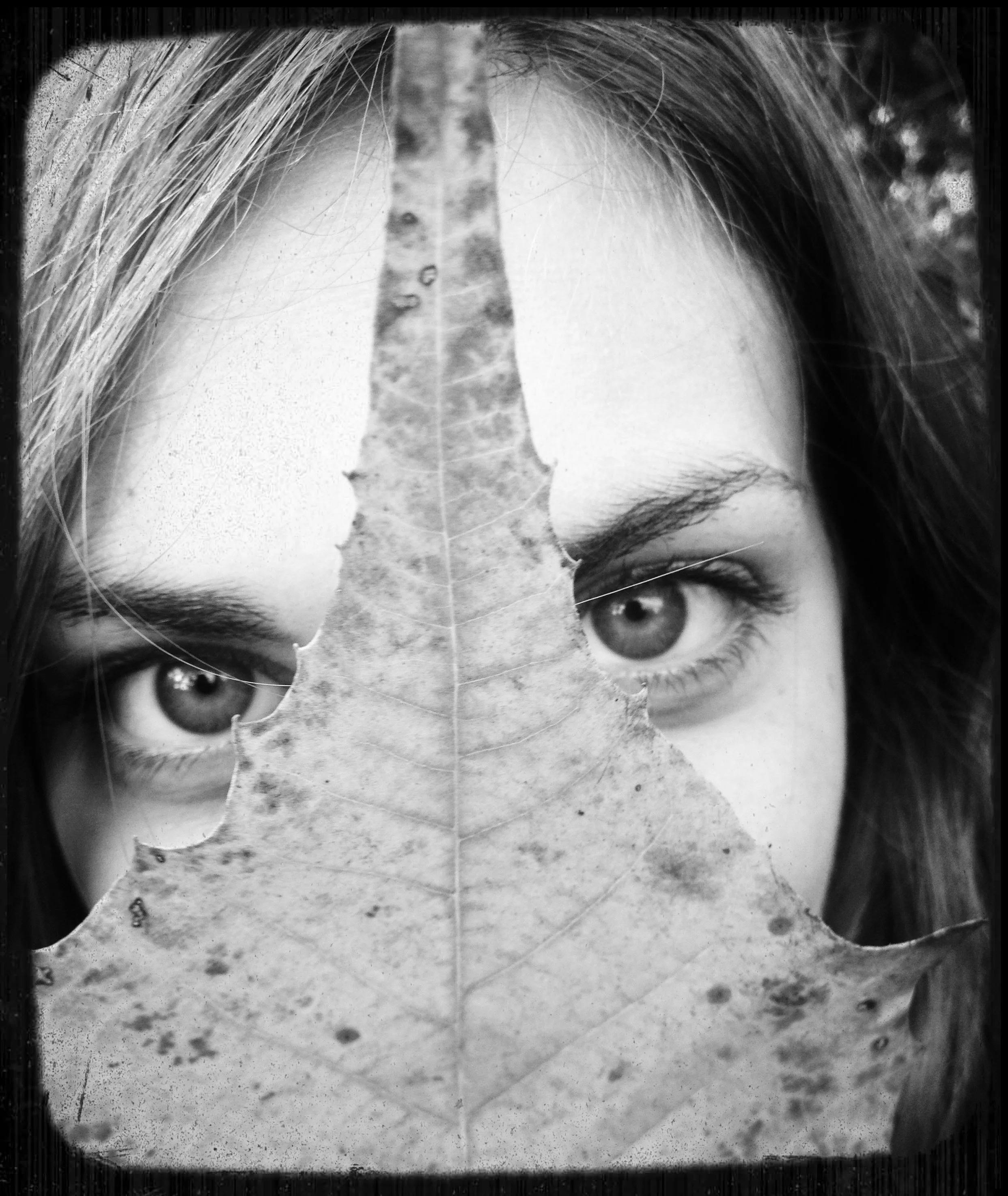 A close-up black and white photo of a person's face, partially obscured by a large leaf, with visible eyes and hair.
