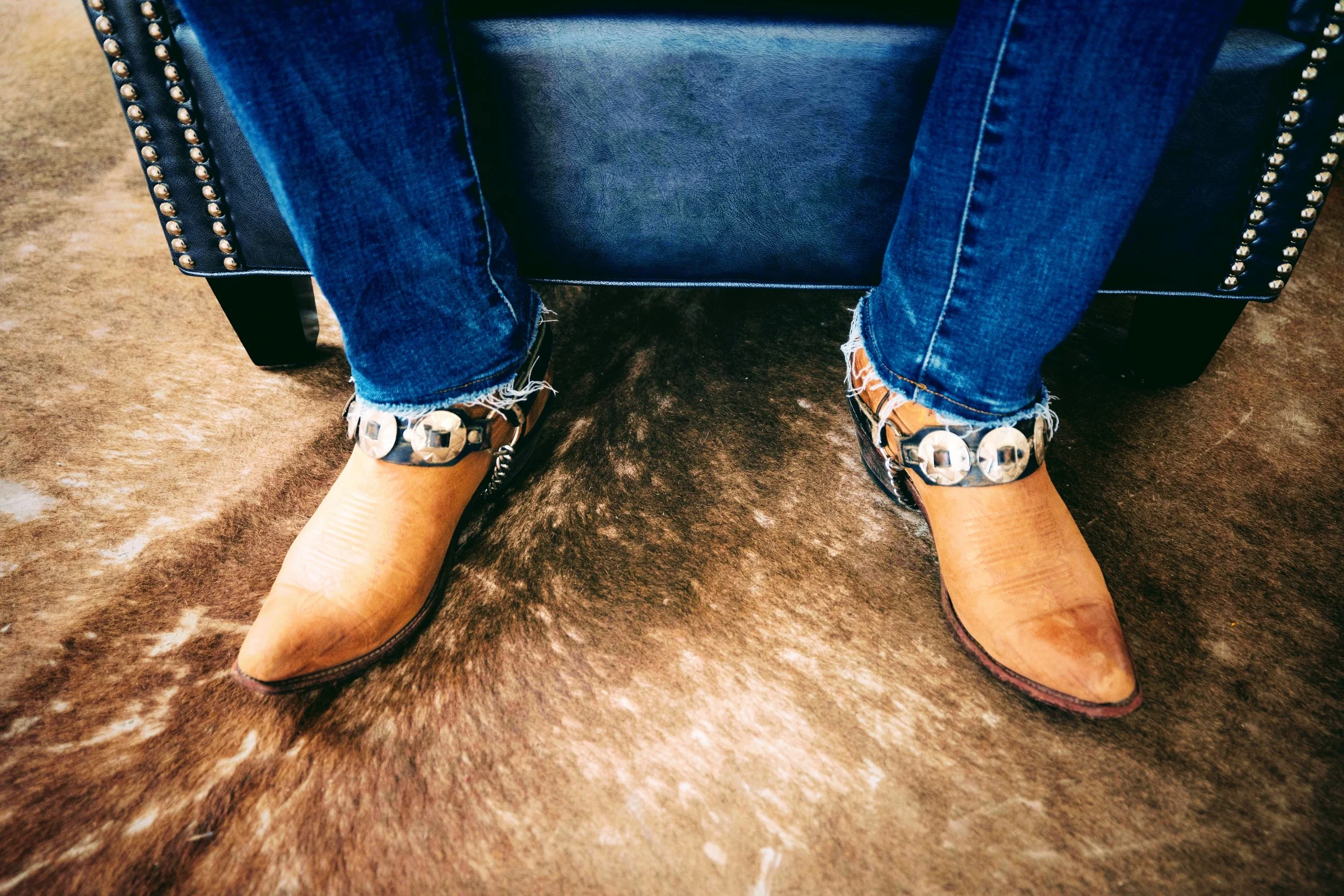 Person sitting on a black leather chair with studded detail, wearing distressed blue jeans and tan cowboy boots with decorative silver accents, on a brown textured carpet.