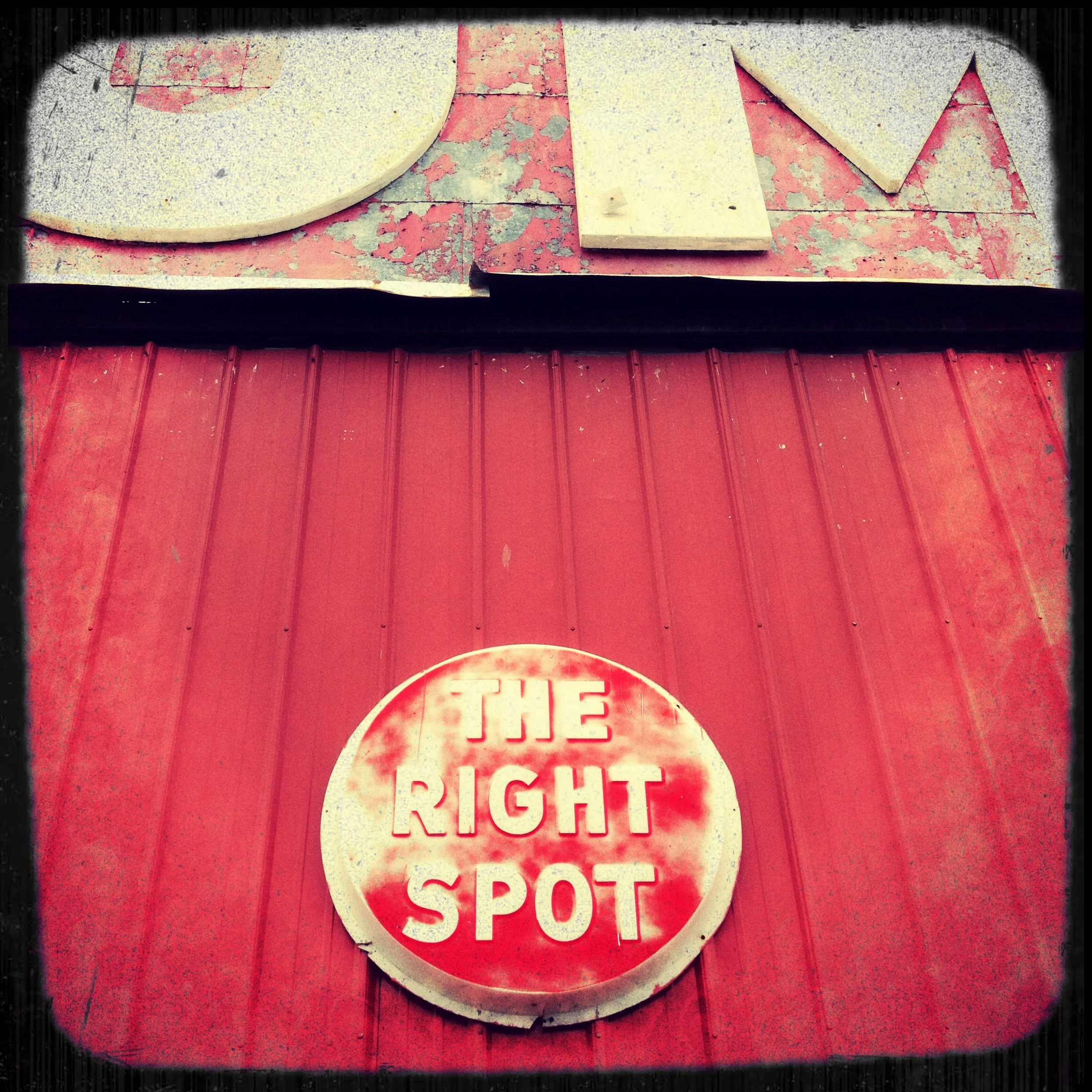 A red circular sign with white outlined text that says "THE RIGHT SPOT" on a red background, attached to a red metal building exterior.