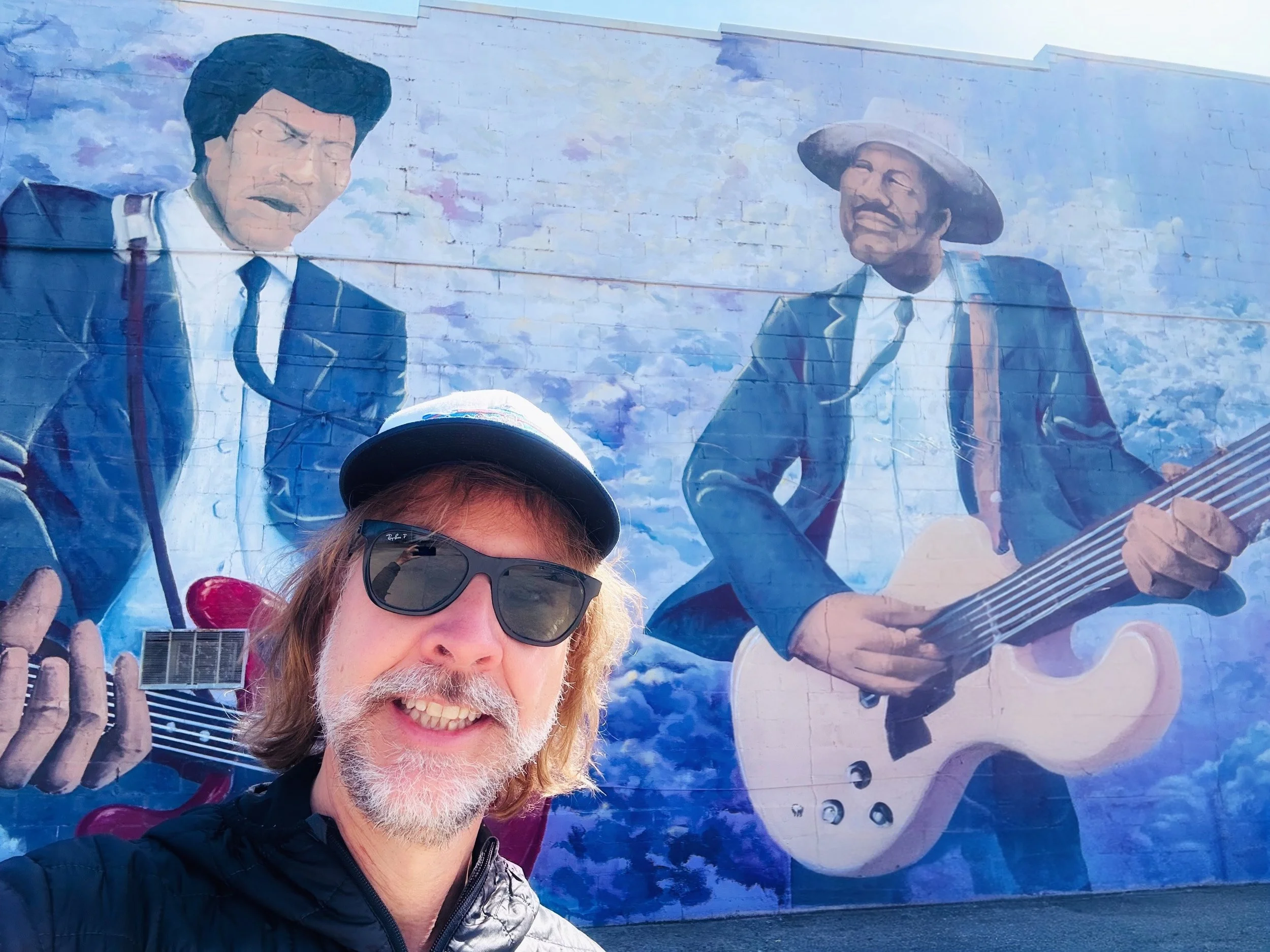 A man with sunglasses, a hat, and a beard taking a selfie in front of a large mural. The mural features two men in suits playing guitars, painted on a brick wall, with one man playing a white guitar and the other a red guitar.