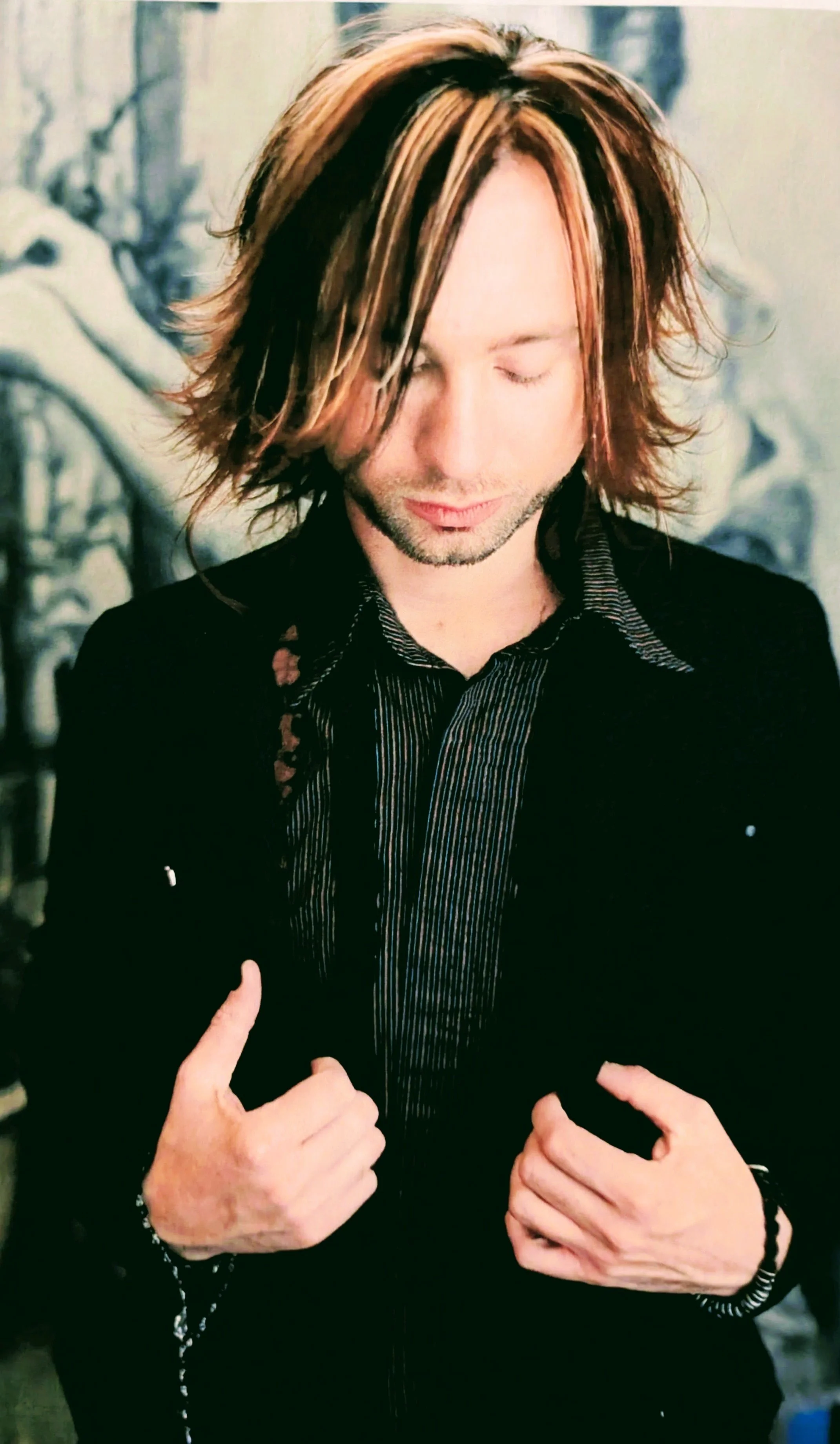 A man with shoulder-length hair with blonde highlights, wearing a black jacket and dark vertically striped shirt, is making a thumbs-up gesture and smiling with eyes closed. The background appears to be an outdoor winter scene with snow-covered trees