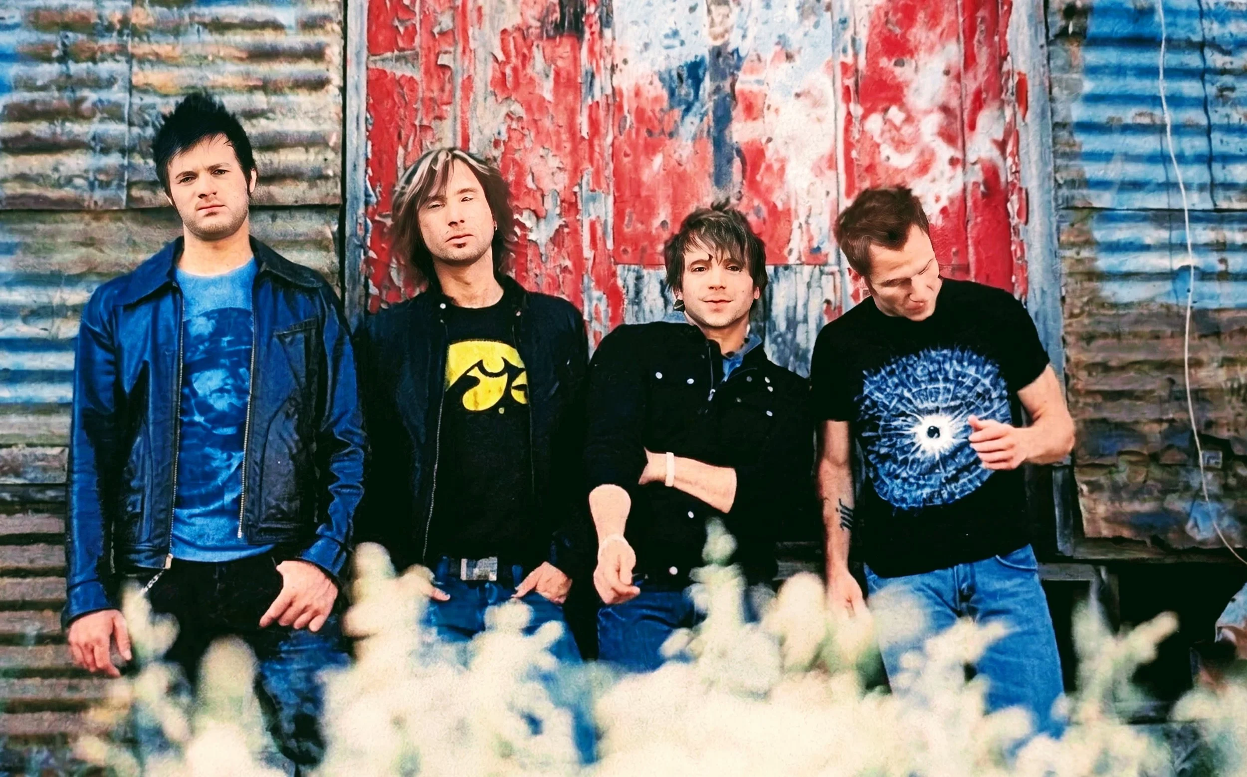 Four men standing in front of a weathered, graffiti-covered wall, with white flowers at the bottom of the image.