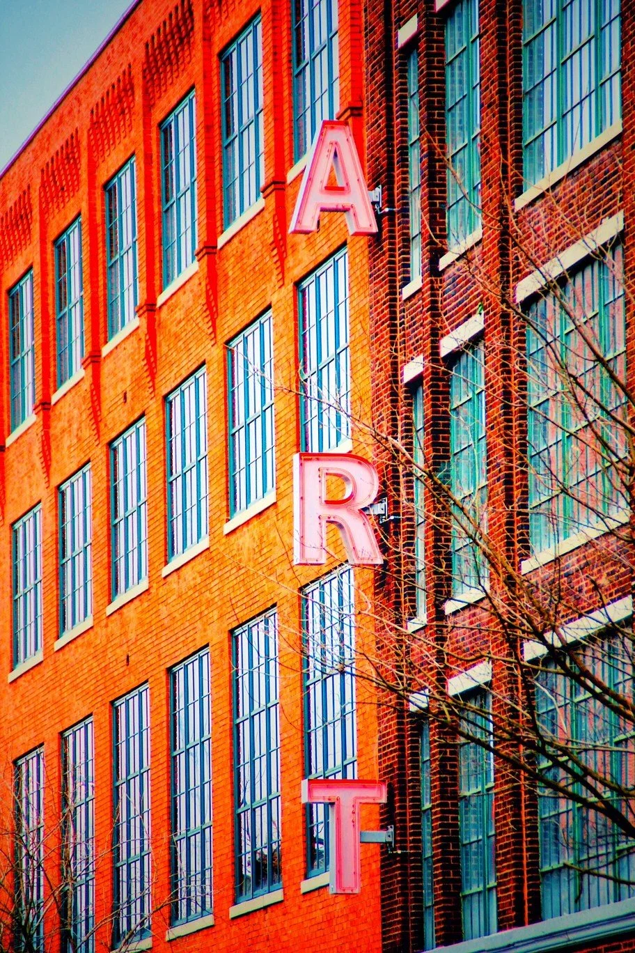 Neon pink and white sign spelling 'ART' on the exterior of a red brick building with tall windows, some bare tree branches in the foreground.