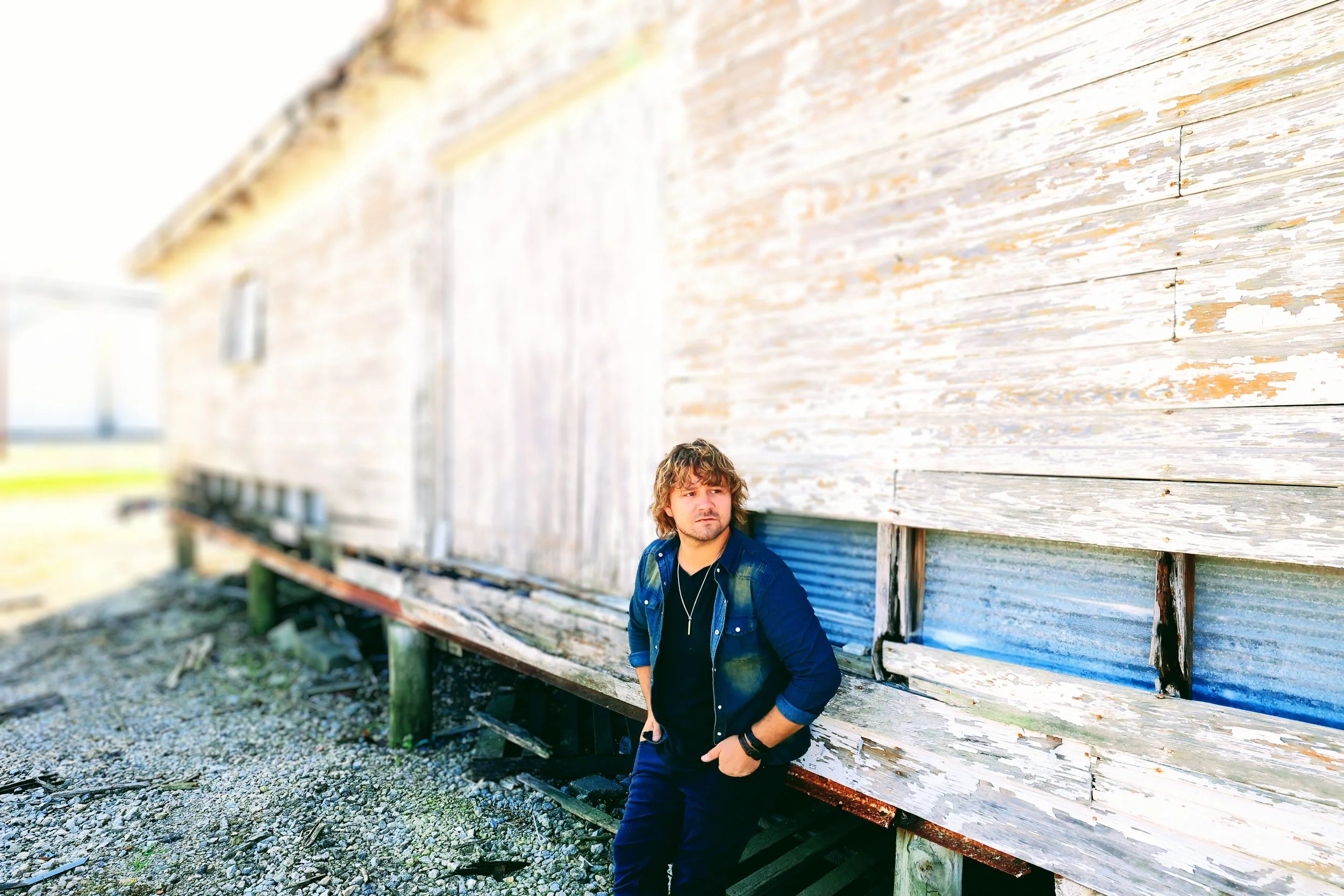 A young man with curly hair and casual clothing leaning against an old weathered building with peeling paint.