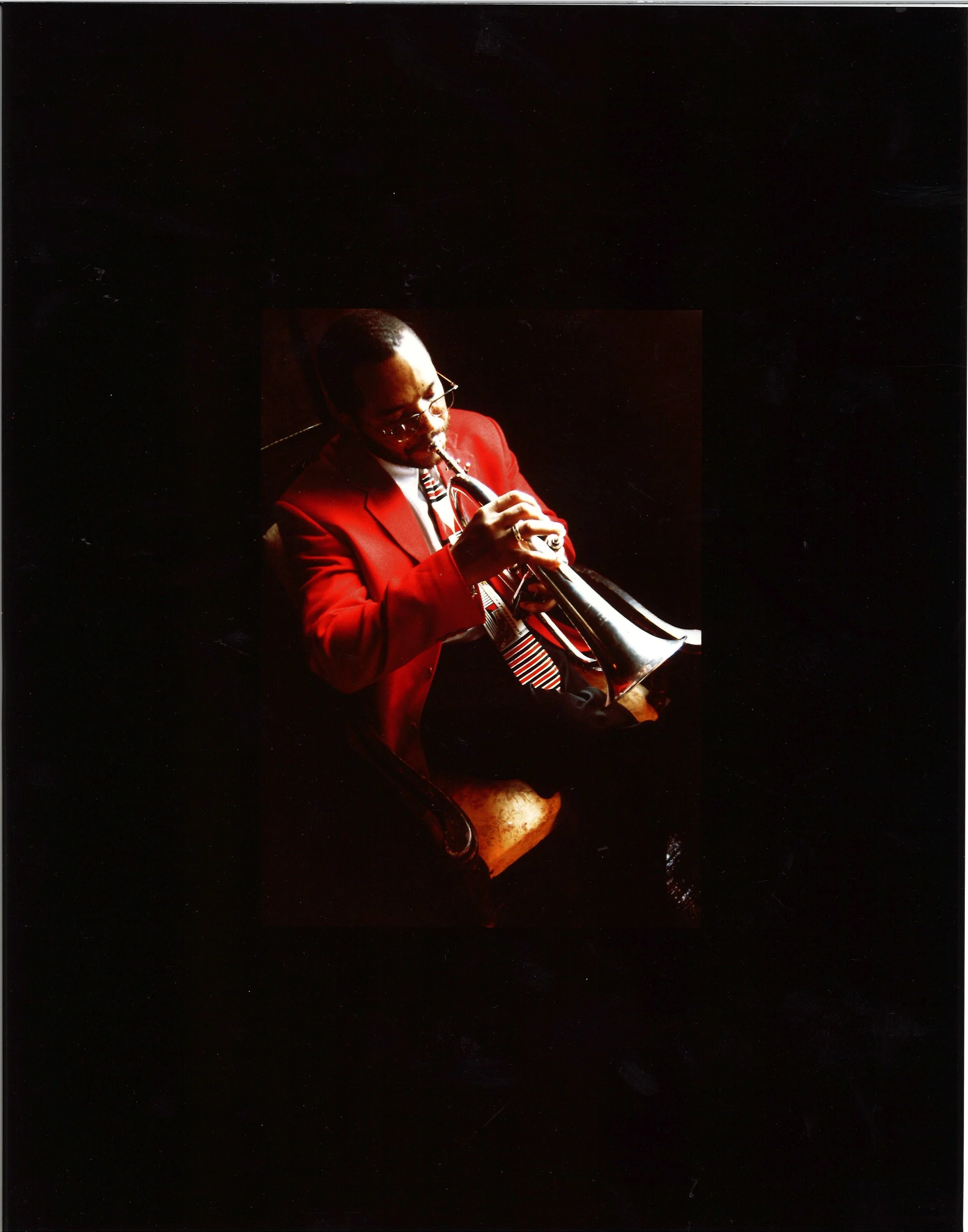 Man in a red blazer playing a trumpet, sitting on a vintage chair, with a dark background.