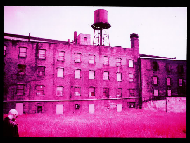 An old brick building with multiple windows, a water tower on top, and a grassy foreground bathed in pink light.