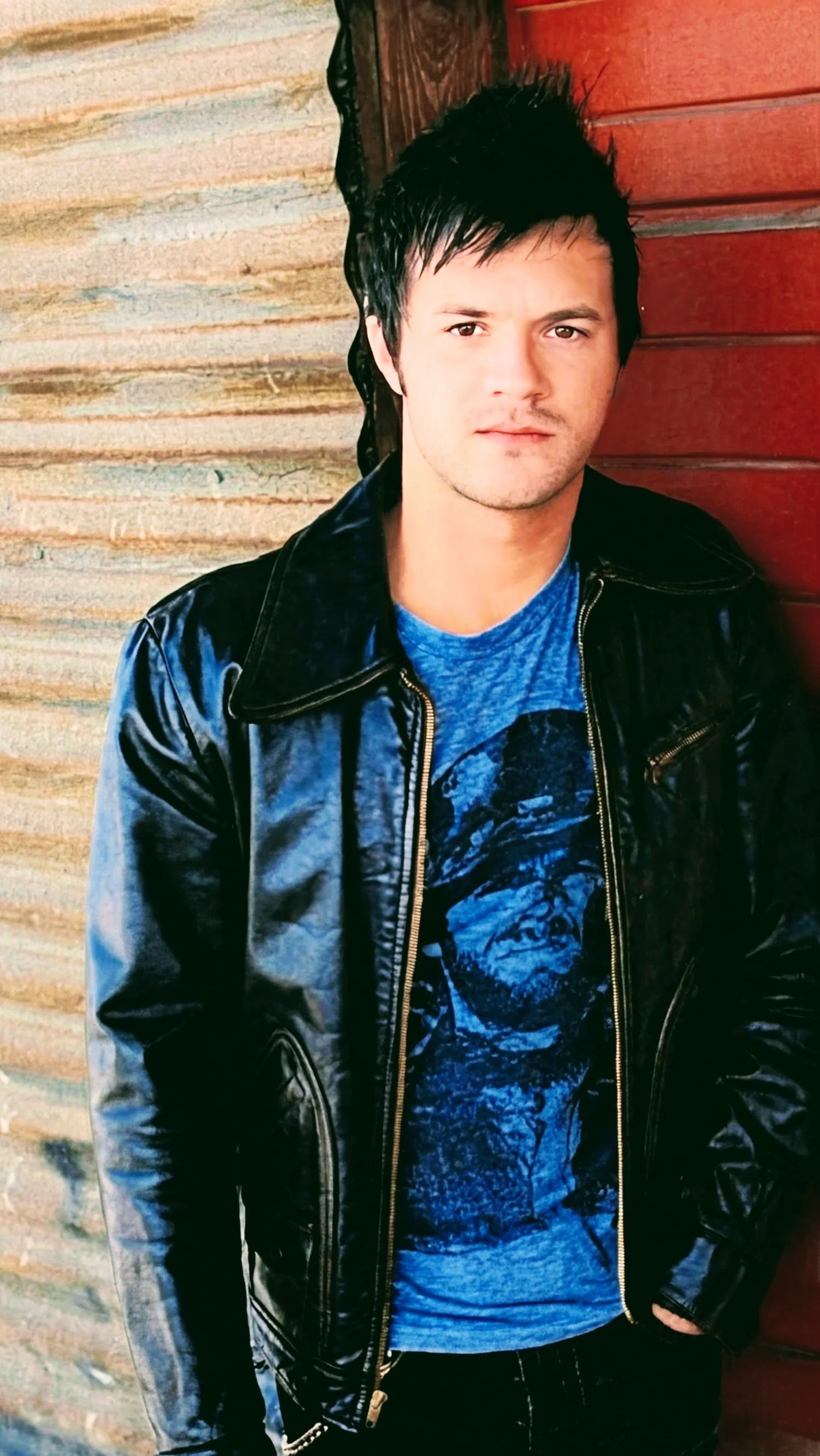 Young man with dark hair, wearing a black leather jacket and a blue graphic t-shirt, standing against a rustic wooden and brick wall.