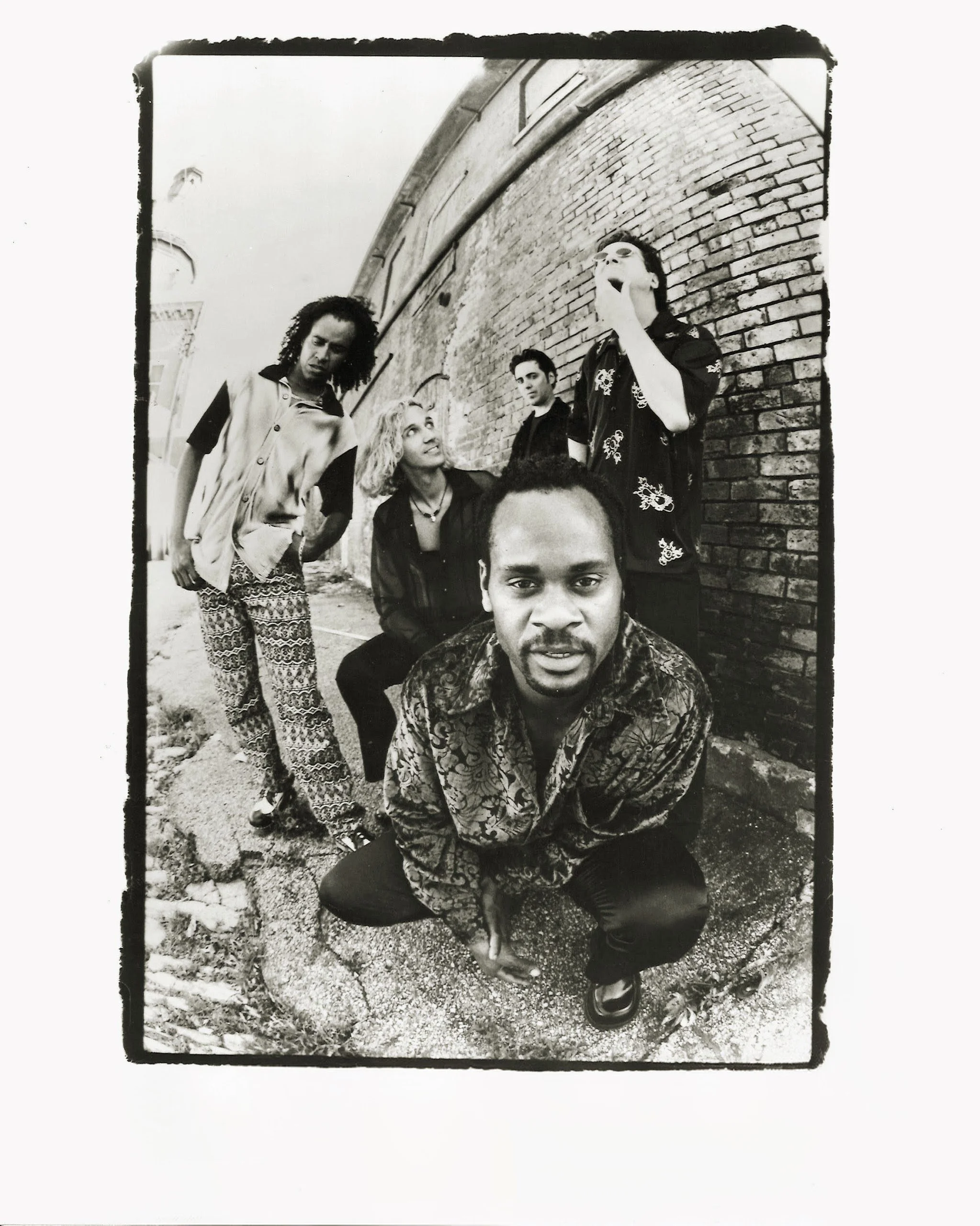 A black and white photo of five diverse young adults standing and sitting against a brick wall outside, with one person in the group showing an expressive gesture.