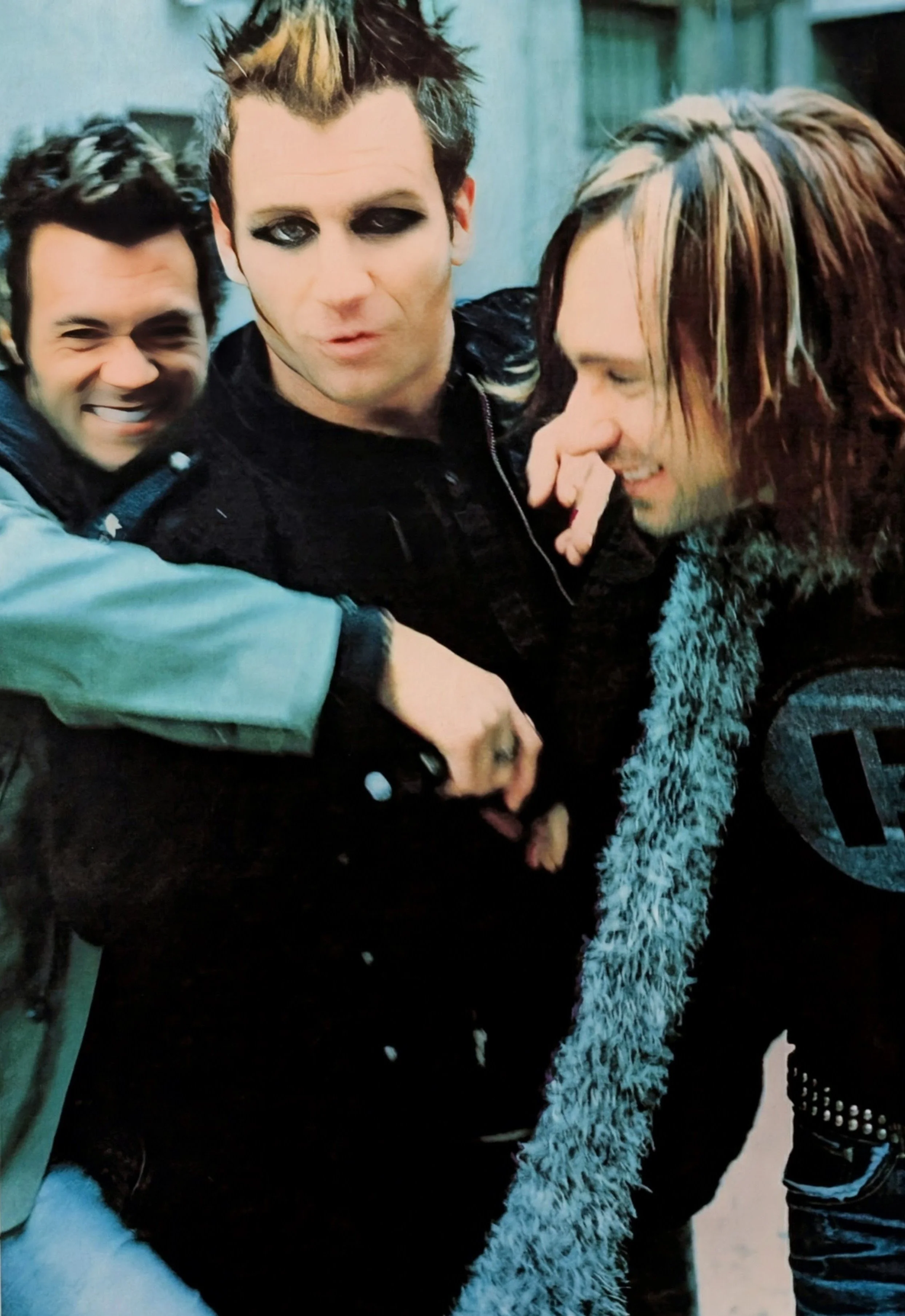 Three men, two with long hair and makeup, are huddled closely together, with one man in the middle looking directly at the camera. They are all smiling or making playful expressions.