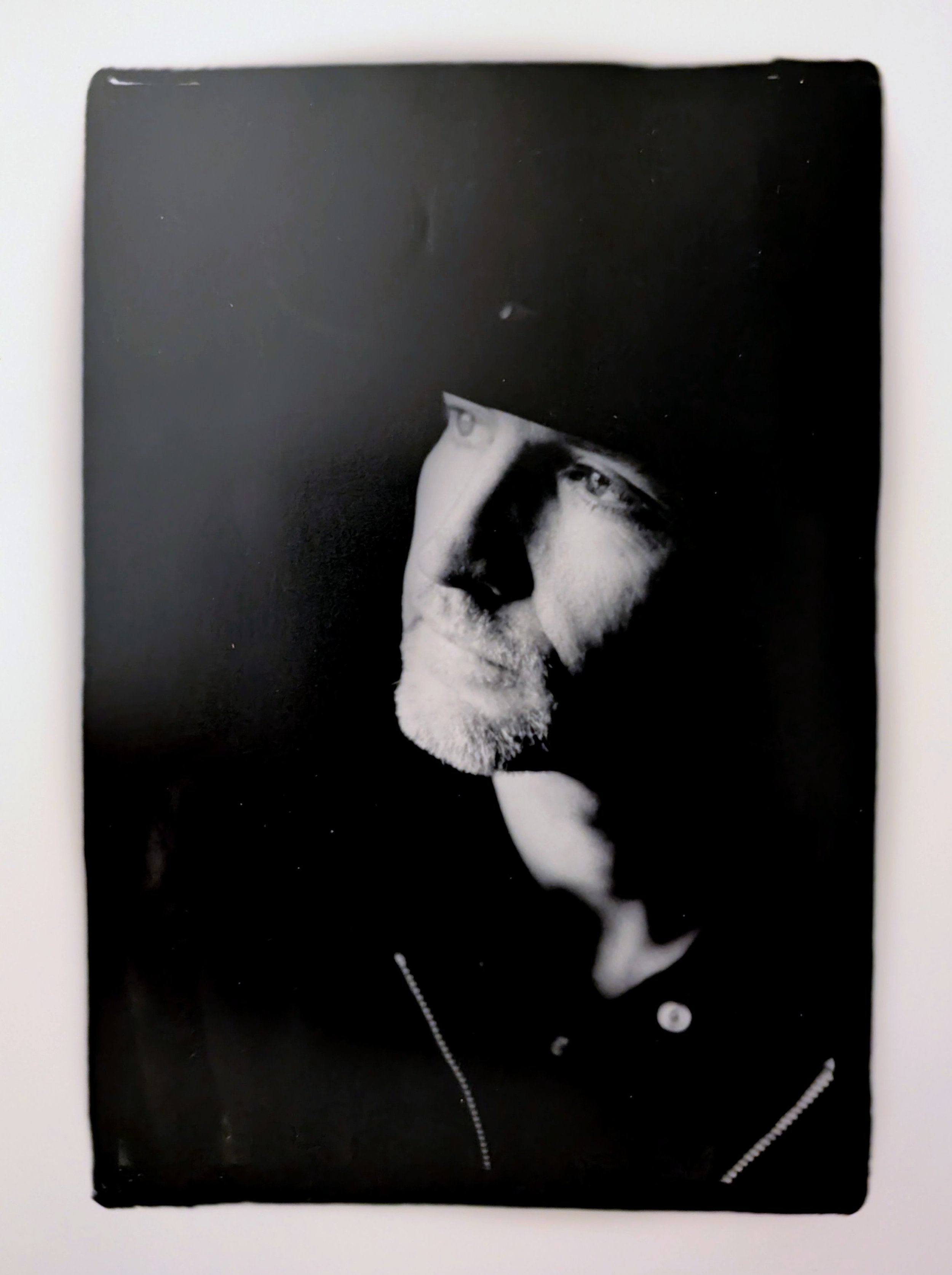 Black and white portrait photograph of a man with a beard and mustache, wearing a black coat with zippers, looking contemplative to the side.