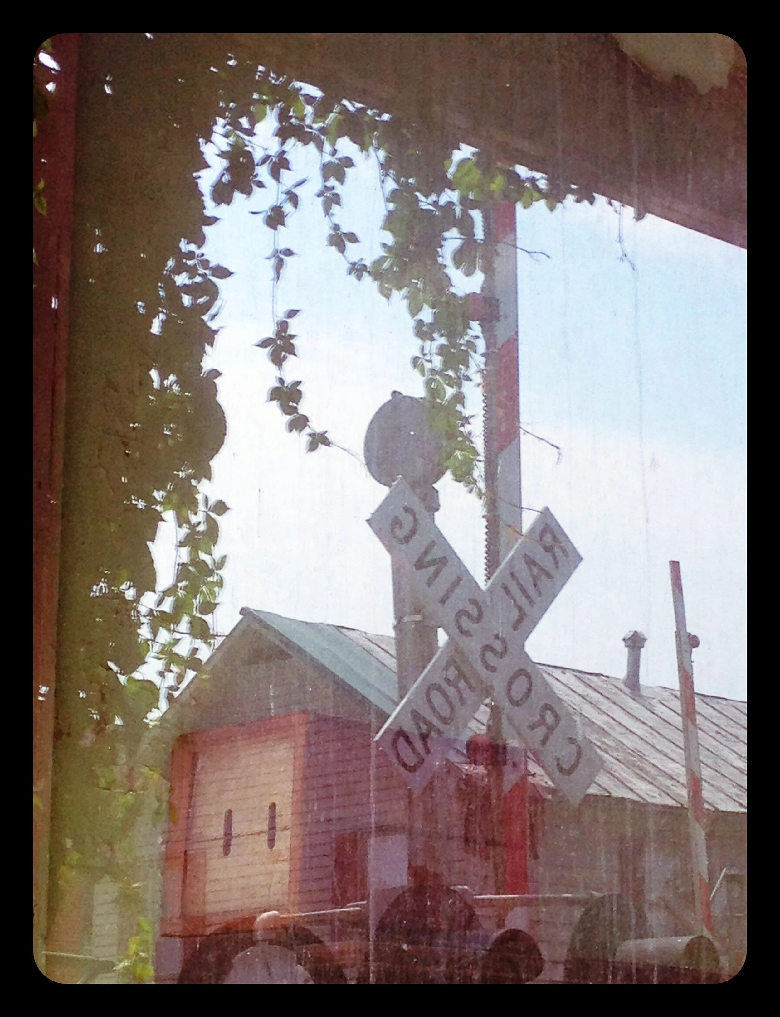A weathered railroad crossing sign reads 'RAILROAD CROSSING'. The sign is mounted on a pole with a background of an old building with a metal roof, trees, and a cloudy sky, as seen through a glass window.