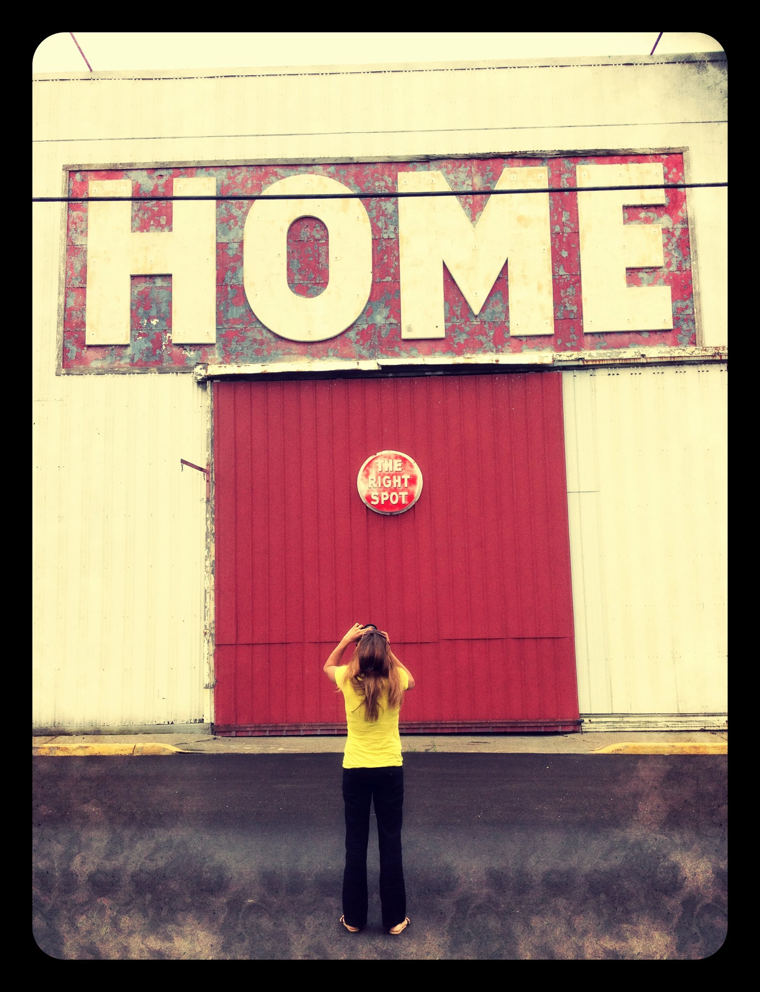 A woman in a yellow shirt and black pants standing in front of a large red metal door with a sign that reads 'THE RIGHT SPOT'. Above the door is a sign with the word 'HOME' in large, faded white letters on a red background.