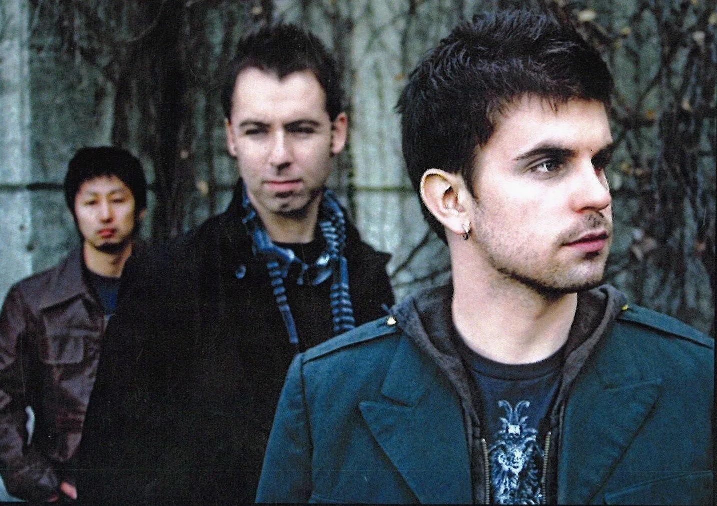 Three young men outdoors in front of trees and stone columns, with the man closest to the camera wearing an earring and a dark jacket.