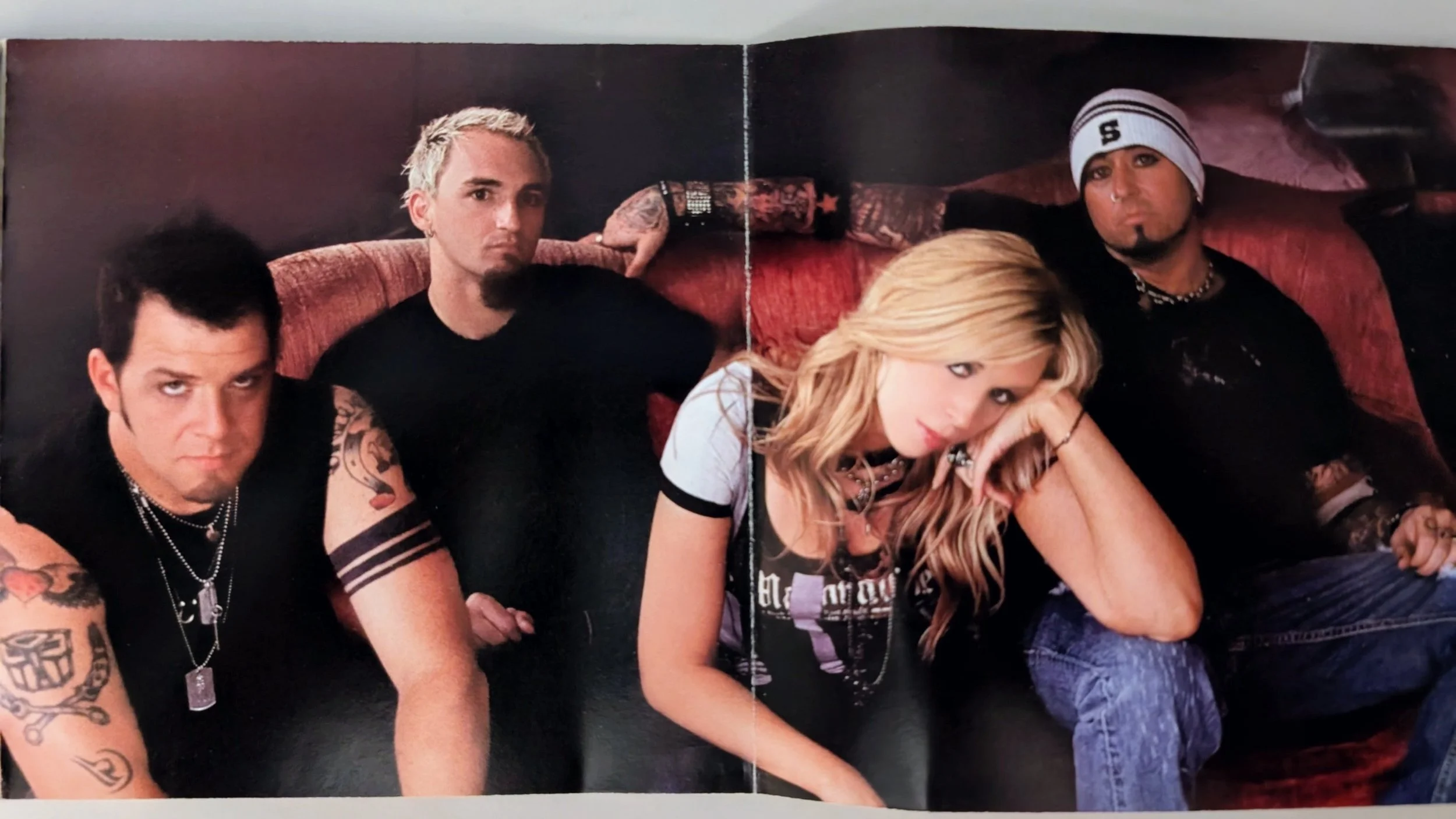 A group of five young adults sitting on a red couch against a dark wall. They are dressed in casual, rock-inspired clothing with tattoos and accessories.