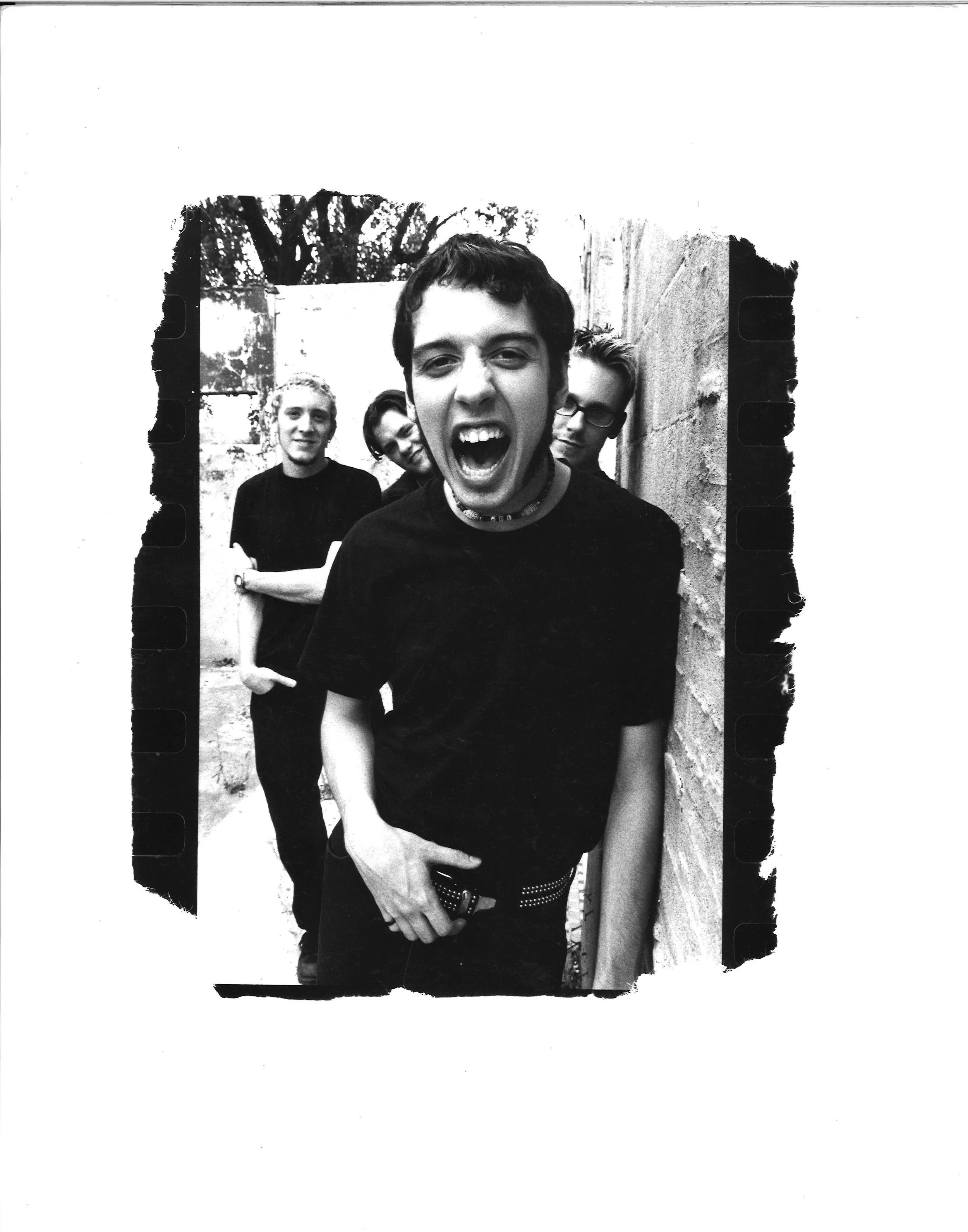 Black and white photo of four young men standing outdoors, with one in the foreground showing an open mouth, wearing a choker and a black t-shirt, while the others pose behind him.