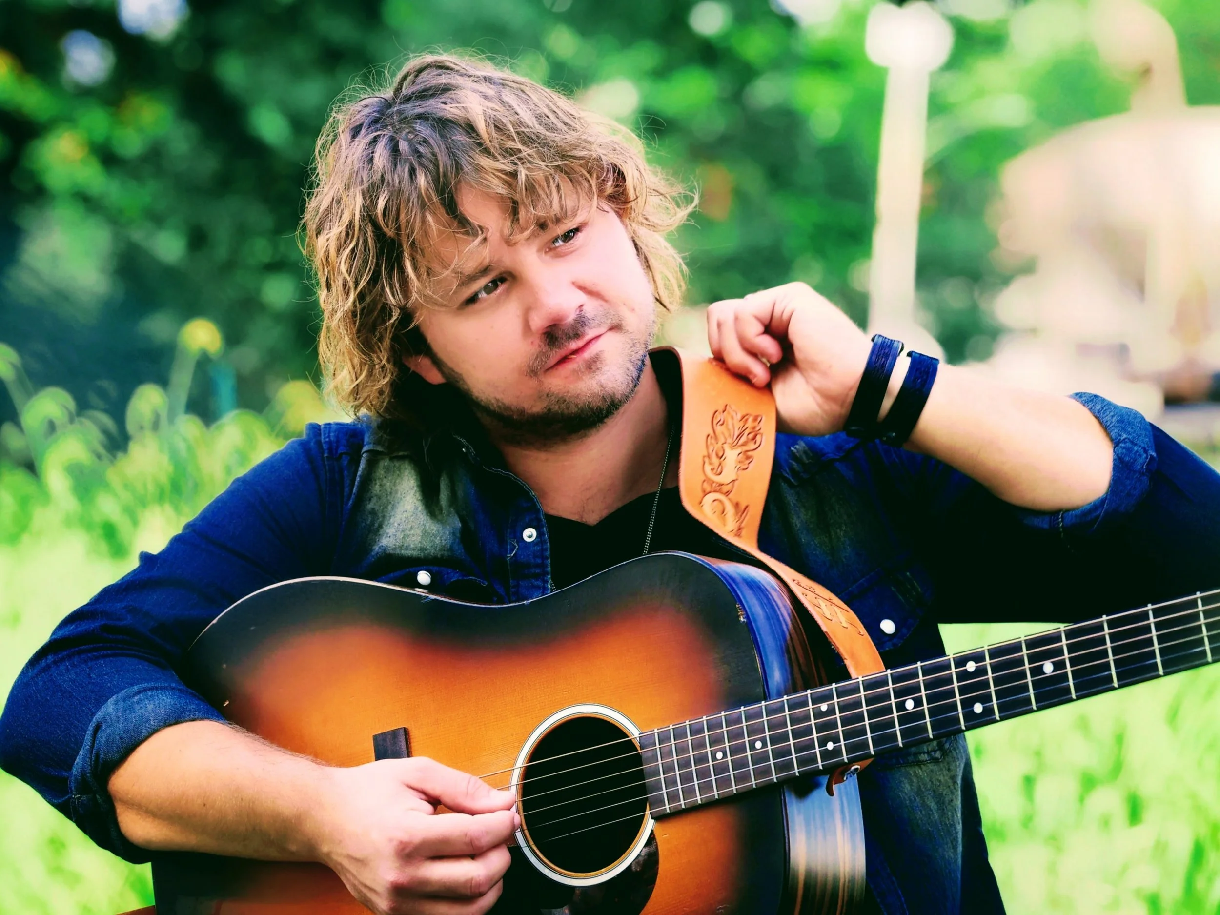 A man with light skin, curly blonde hair, and a beard playing an acoustic guitar outdoors with green trees in the background.