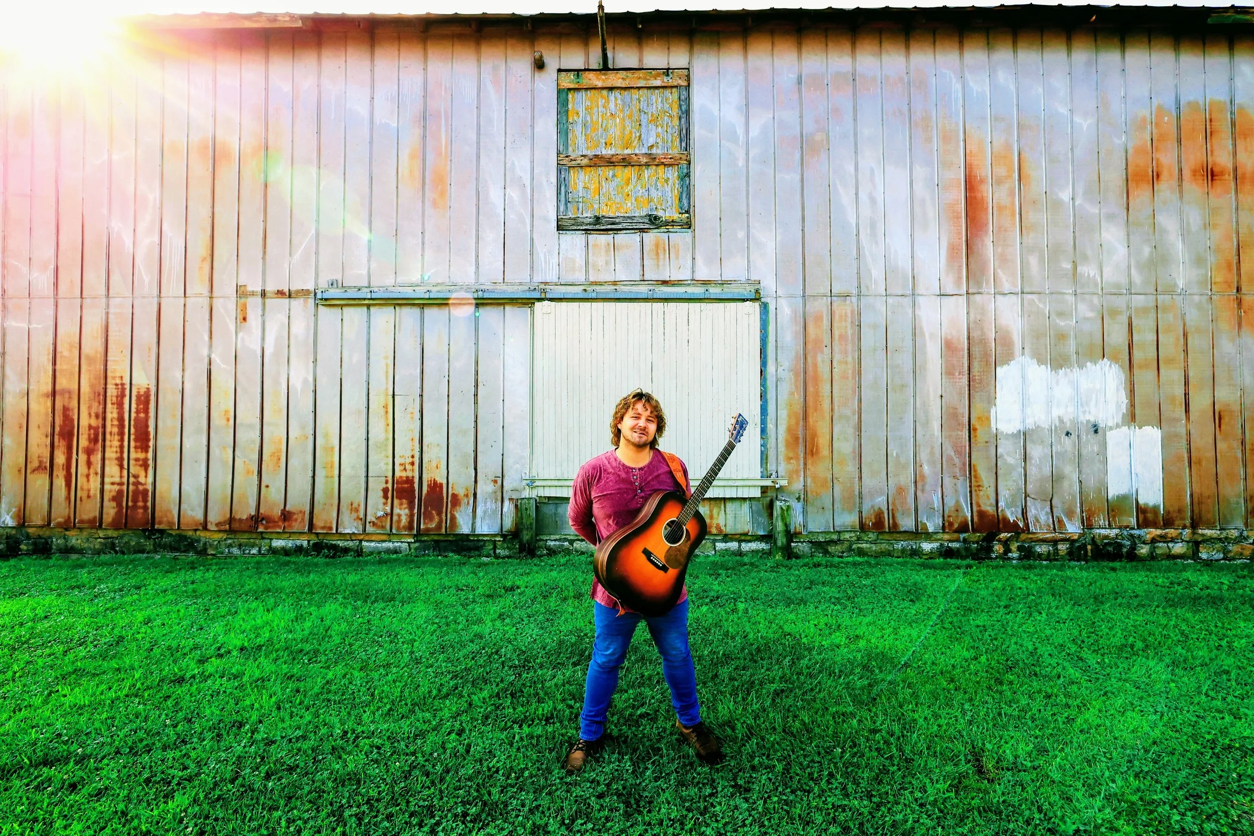 A man with curly hair and a beard, wearing a red long-sleeve shirt and blue jeans, stands on green grass holding an acoustic guitar in front of a large, weathered metal building with a small window and patches of peeling paint. The sunlight creates a