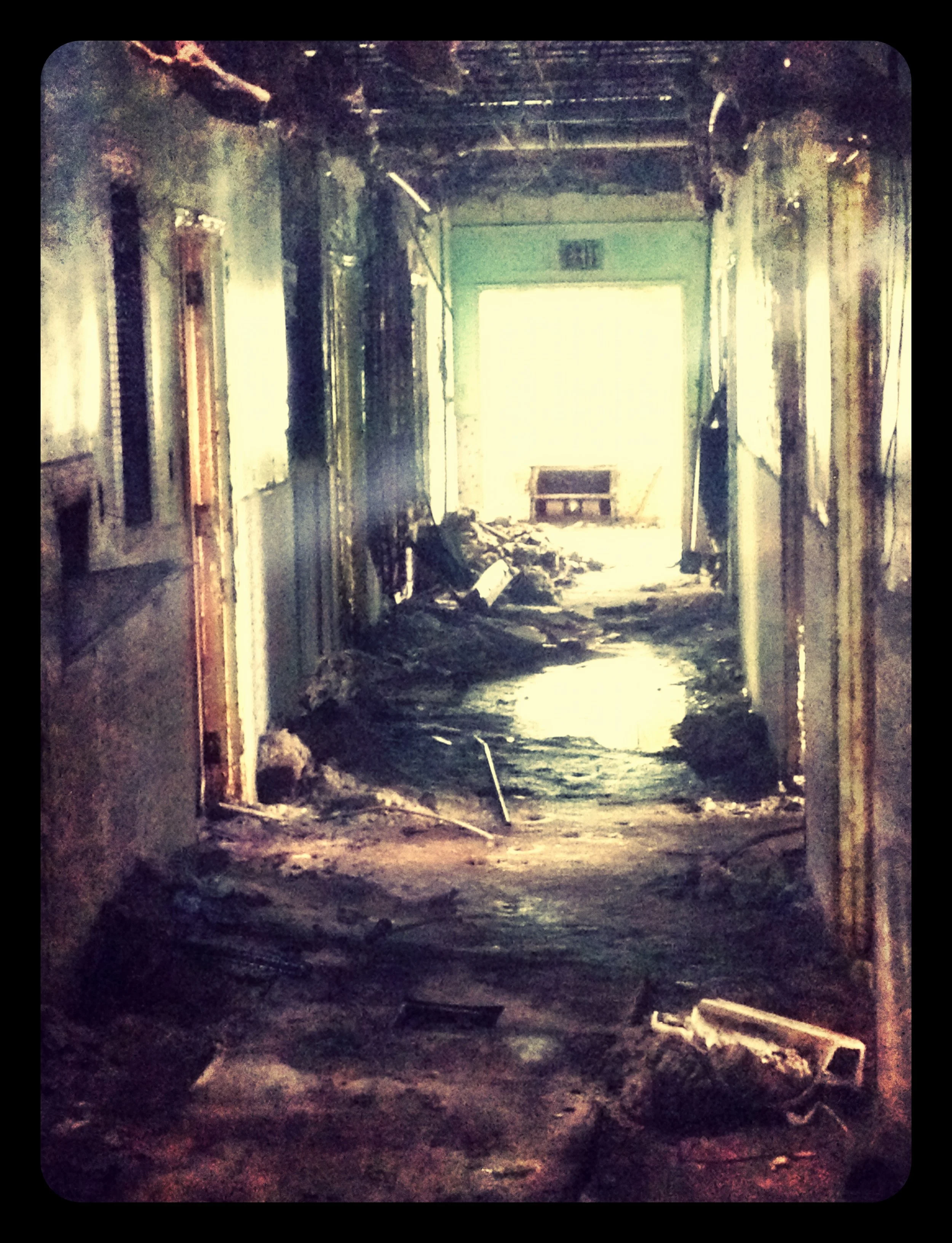 Interior of a heavily damaged and burnt building with debris scattered on the floor, charred walls, and soot stains, with bright light coming from an opening at the end of the hallway.