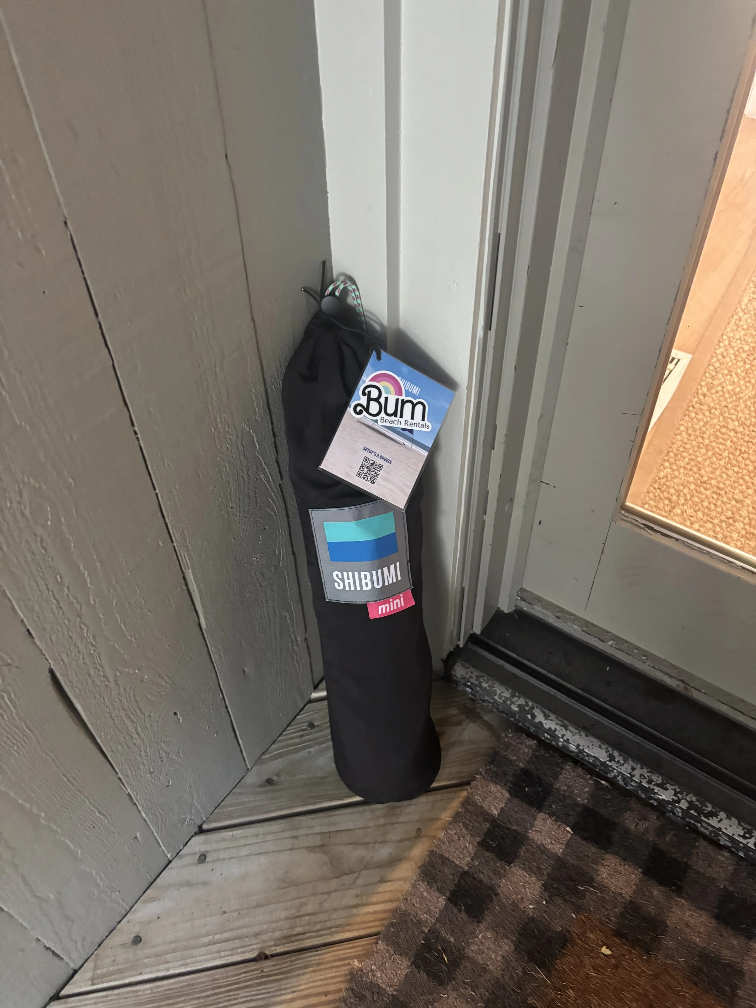 A black duffel bag with a tag labeled 'SHIBUMI mini' leaning against a wall near a door in an indoor setting with wooden flooring and a carpeted area.