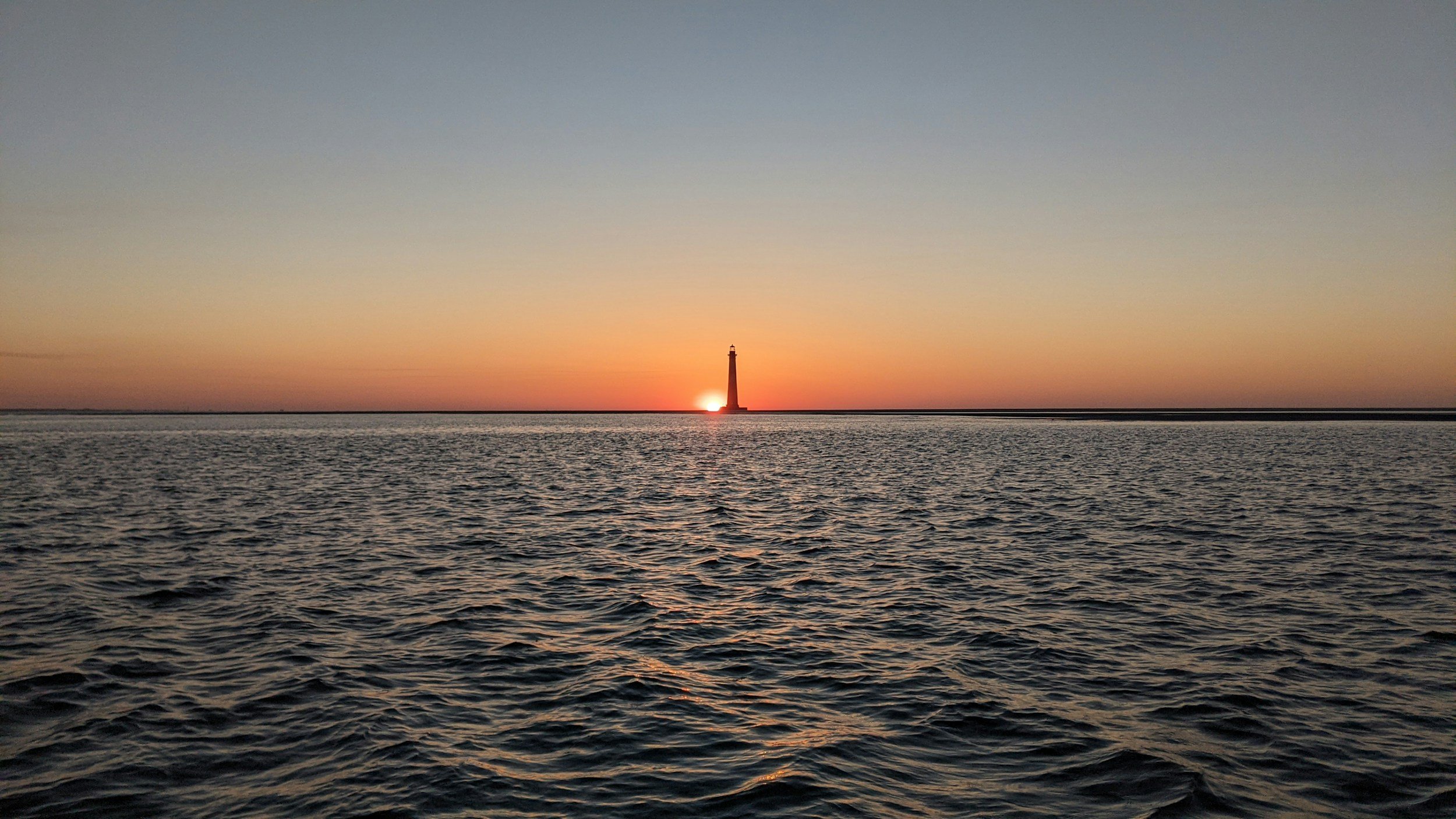 Sunset over the ocean with a lighthouse silhouette in the distance, calm water in the foreground, colorful sky with gradient from blue to orange near the horizon.