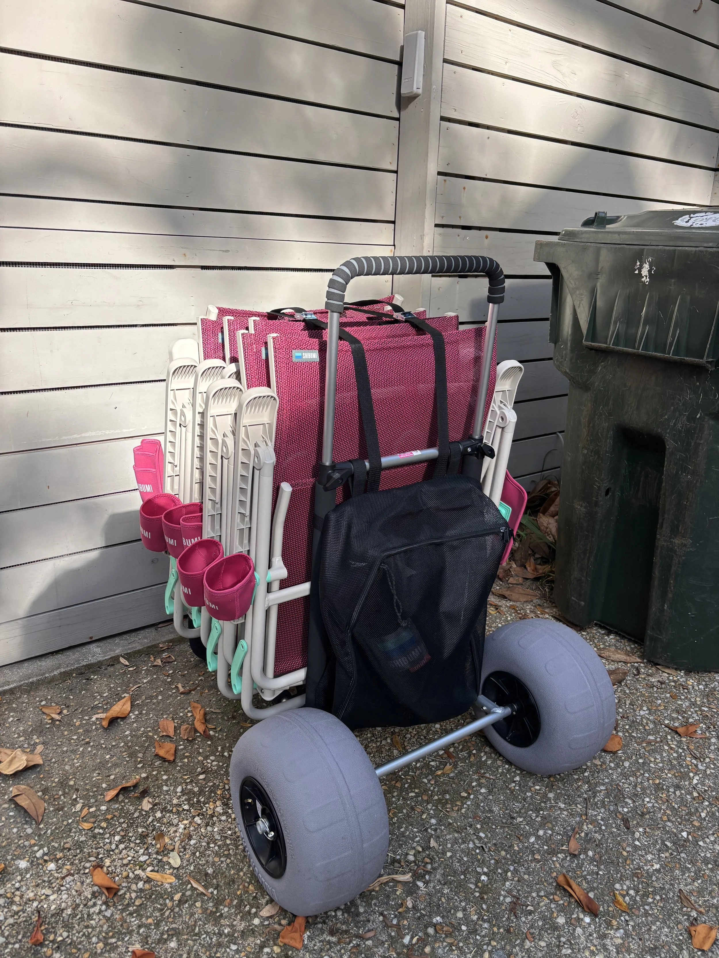 A roller wheel cart with pink and white folding chairs stacked and a black bag hanging from the handle, positioned next to a green trash can.