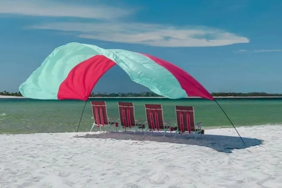 Beach scene with a colorful beach umbrella and five red chairs on white sand, overlooking calm green water under a partly cloudy sky.