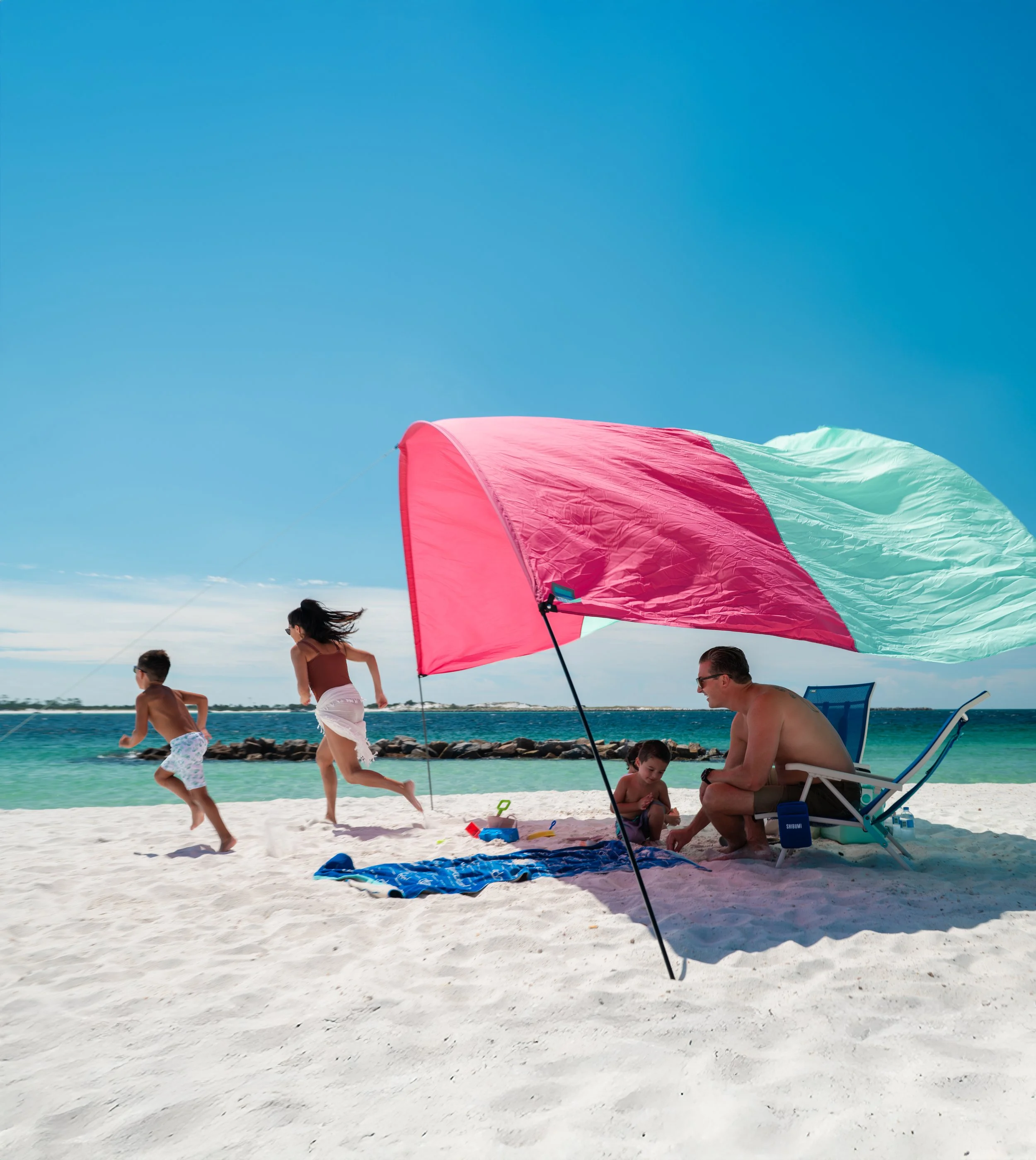 A family enjoying a day at the beach under a pink and blue shibumi shade or umbrella. Two children are running and playing on the sand, while a man and a young boy sit on lounge chairs near the water.