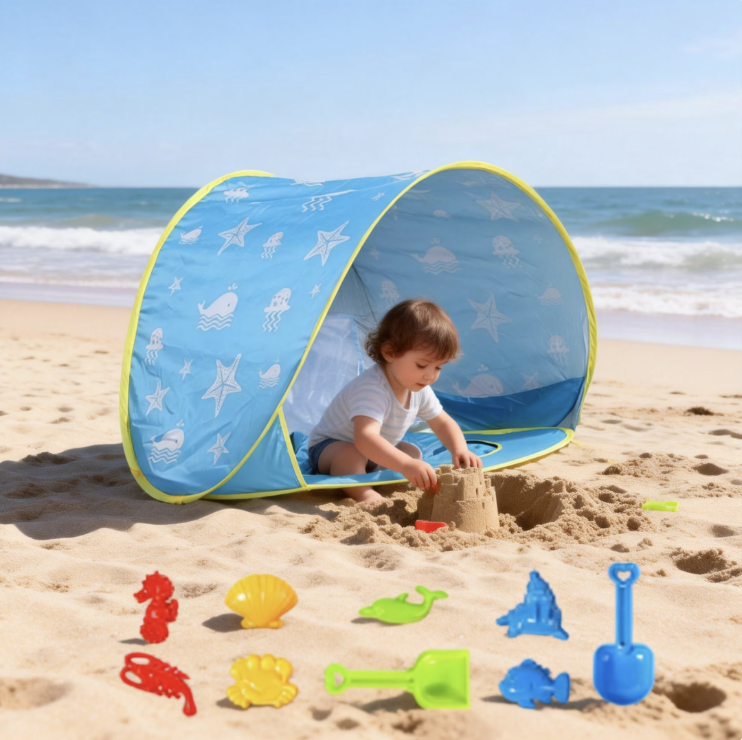 A young child playing with sandcastles and beach toys under a blue sunshade on a sandy beach, with the ocean and blue sky in the background.