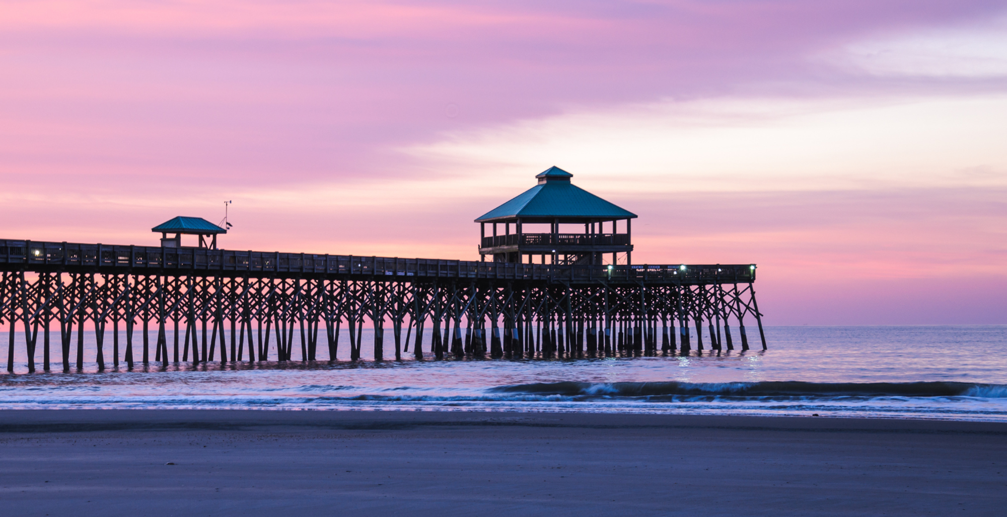 A wooden pier extending over the water at sunset, with a small gazebo-like structure at the end and a larger pavilion. The sky is pink and purple, and gentle waves are seen near the sandy shore.