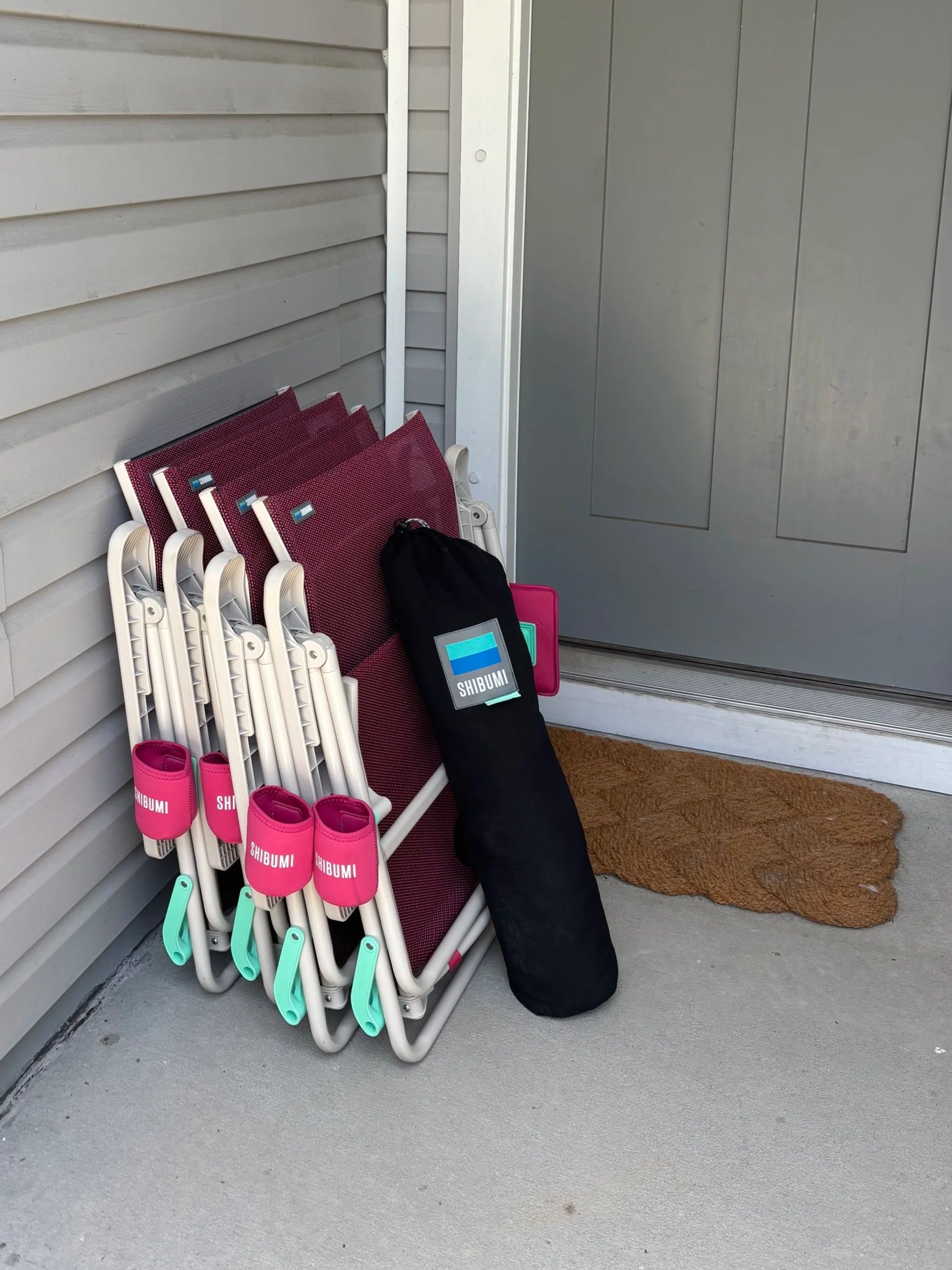 A stack of pink garden tools with white handles and green accents, a black bag with a "SHIBUMI" logo, and a brown doormat outside a house on a concrete porch.