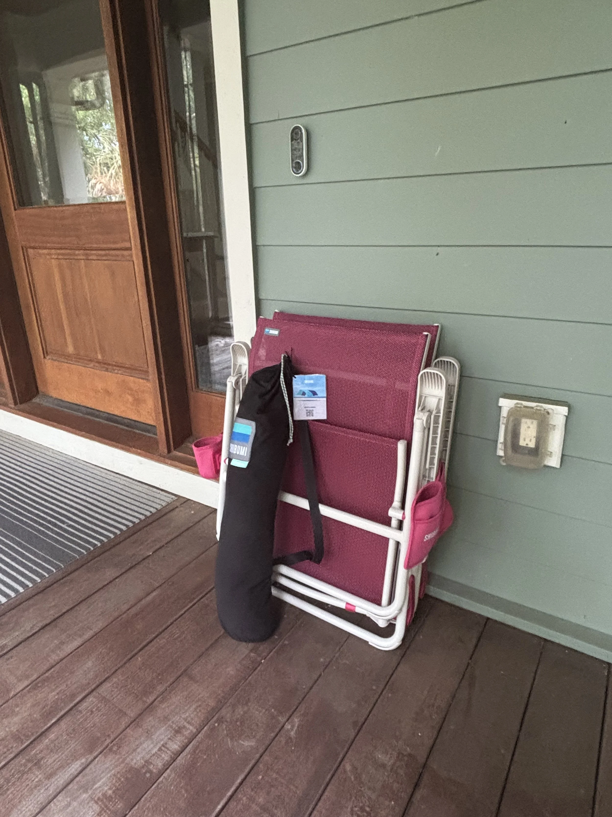 A porch area with wooden flooring, a pink plastic lawn chair with white metal arms, a black backpack and a water bottle on the chair, a pair of pink shoes underneath, and a sealed package inside. The porch has a wooden front door with glass panels, a doorbell, an electrical outlet, and a gray mat at the entrance.