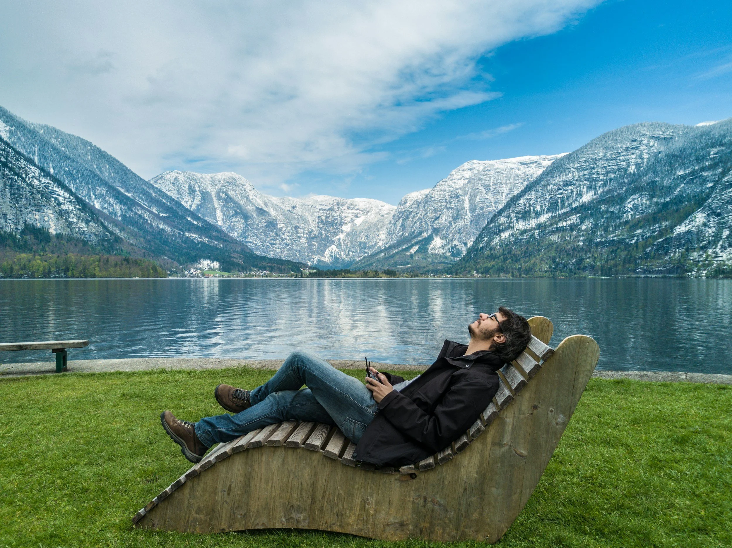 A man relaxing on a wooden bench by a lake with mountains in the background, enjoy the calm and fresh environment.