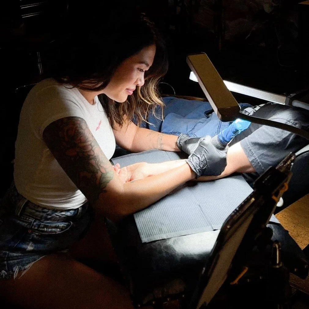 Tattoo artist working on a person's foot with a tattoo machine in a dimly lit studio.